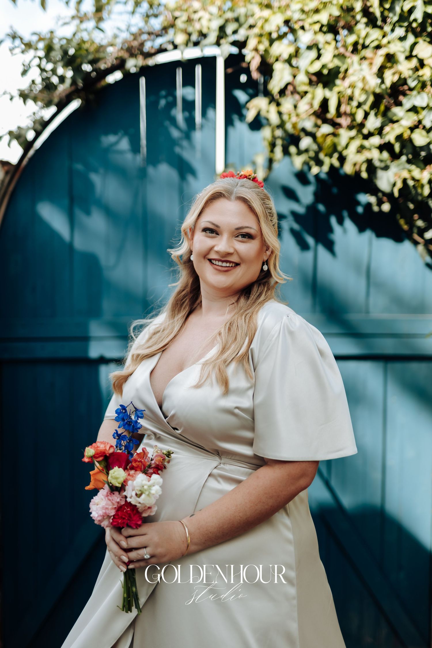 Bride in elegant cream dress with flutter sleeves holds vibrant wildflower bouquet against teal wall backdrop.