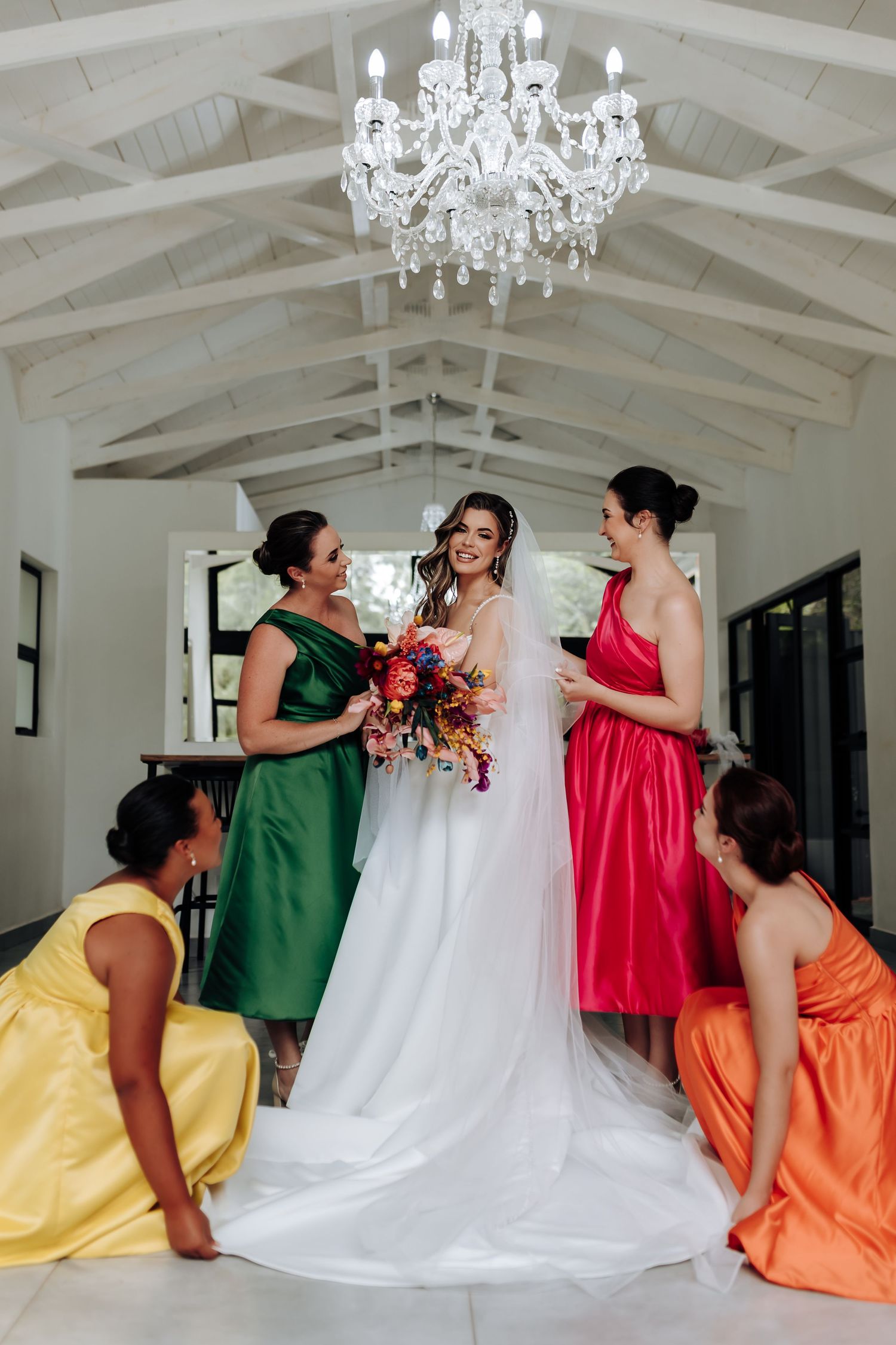 A wedding party in colorful dresses of green, yellow, and coral gather in an elegant white room.