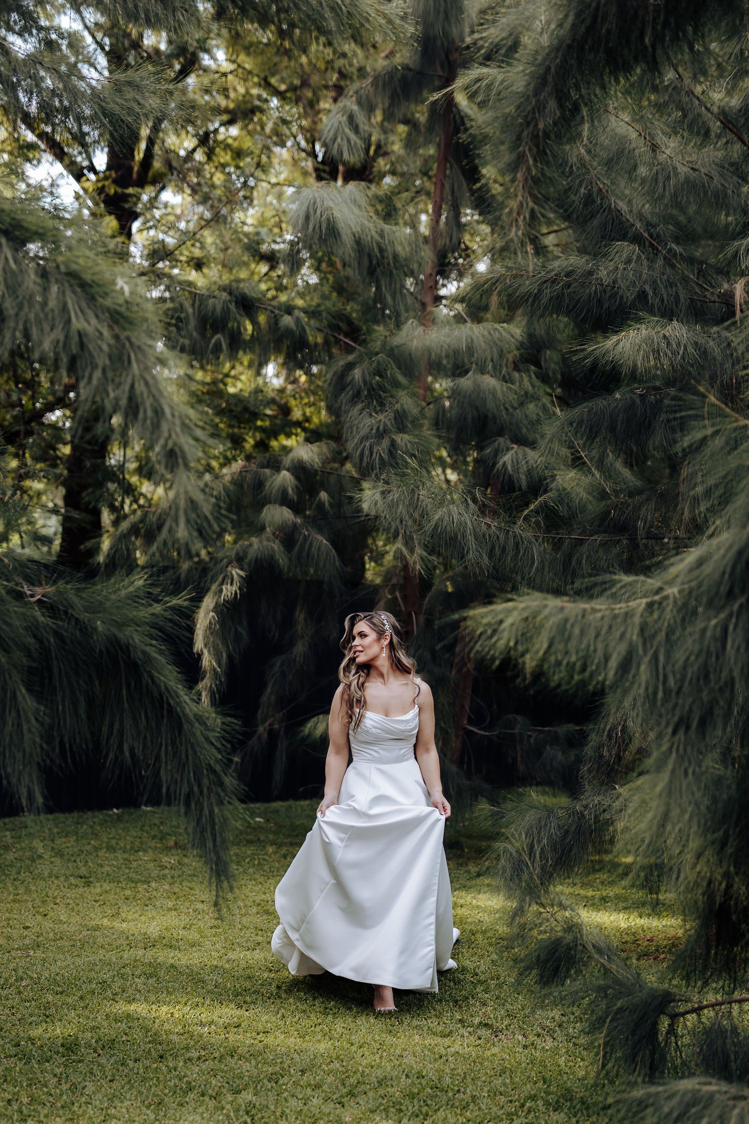 A white dress flows gracefully among evergreen trees in a sunlit garden setting.