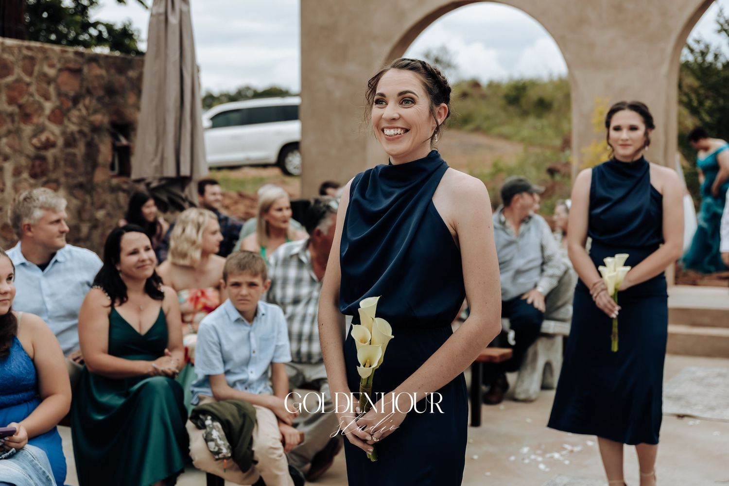 Wedding ceremony guests gather under stone archways at an outdoor venue.
