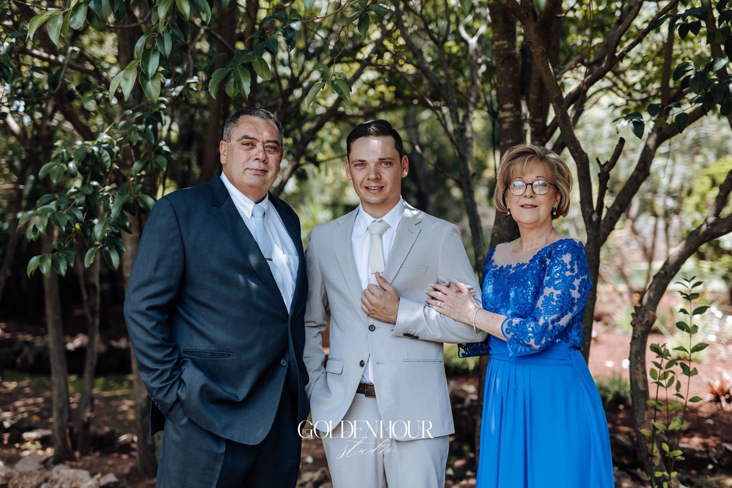 A group of people in formal attire pose together outdoors in a garden setting with lush greenery.