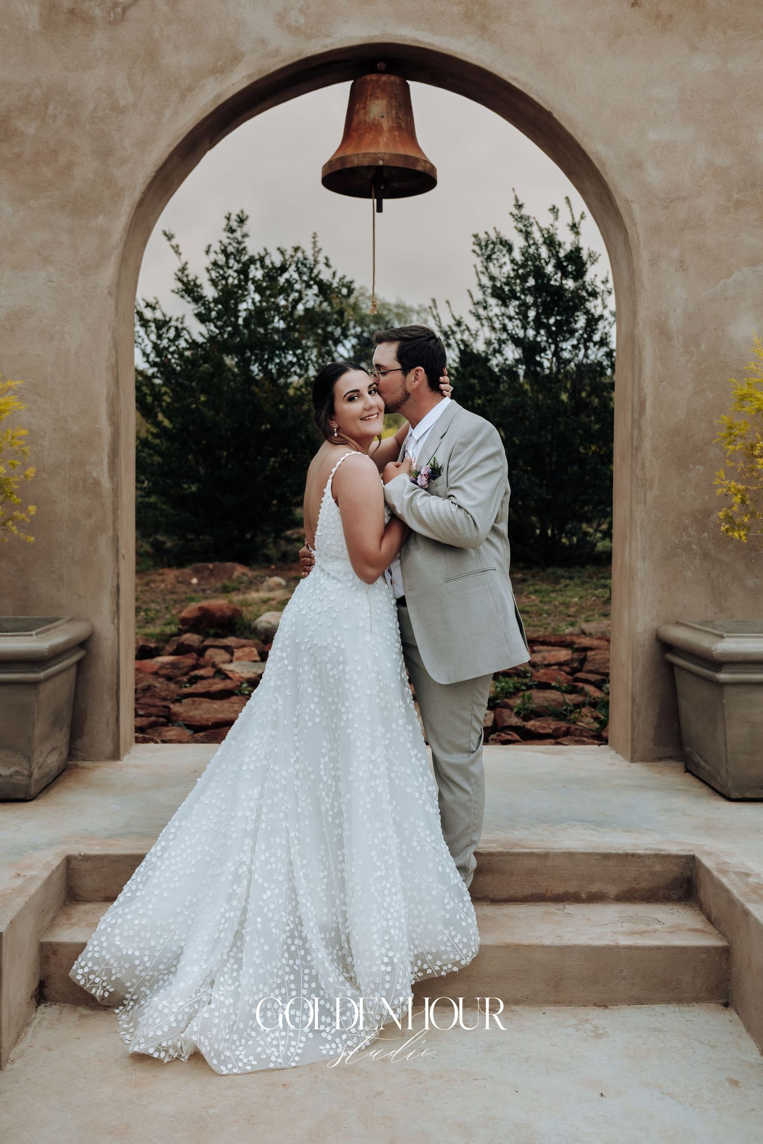 Romantic couple pose under bell-adorned archway on stone steps in elegant wedding attire.