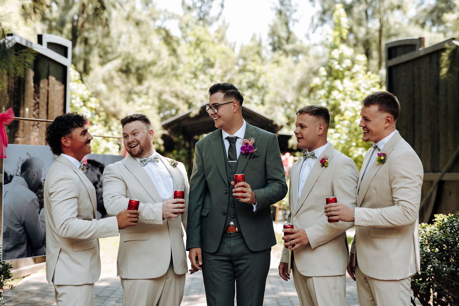 Groomsmen in beige suits and dark suits share drinks and laughs during outdoor wedding celebration.