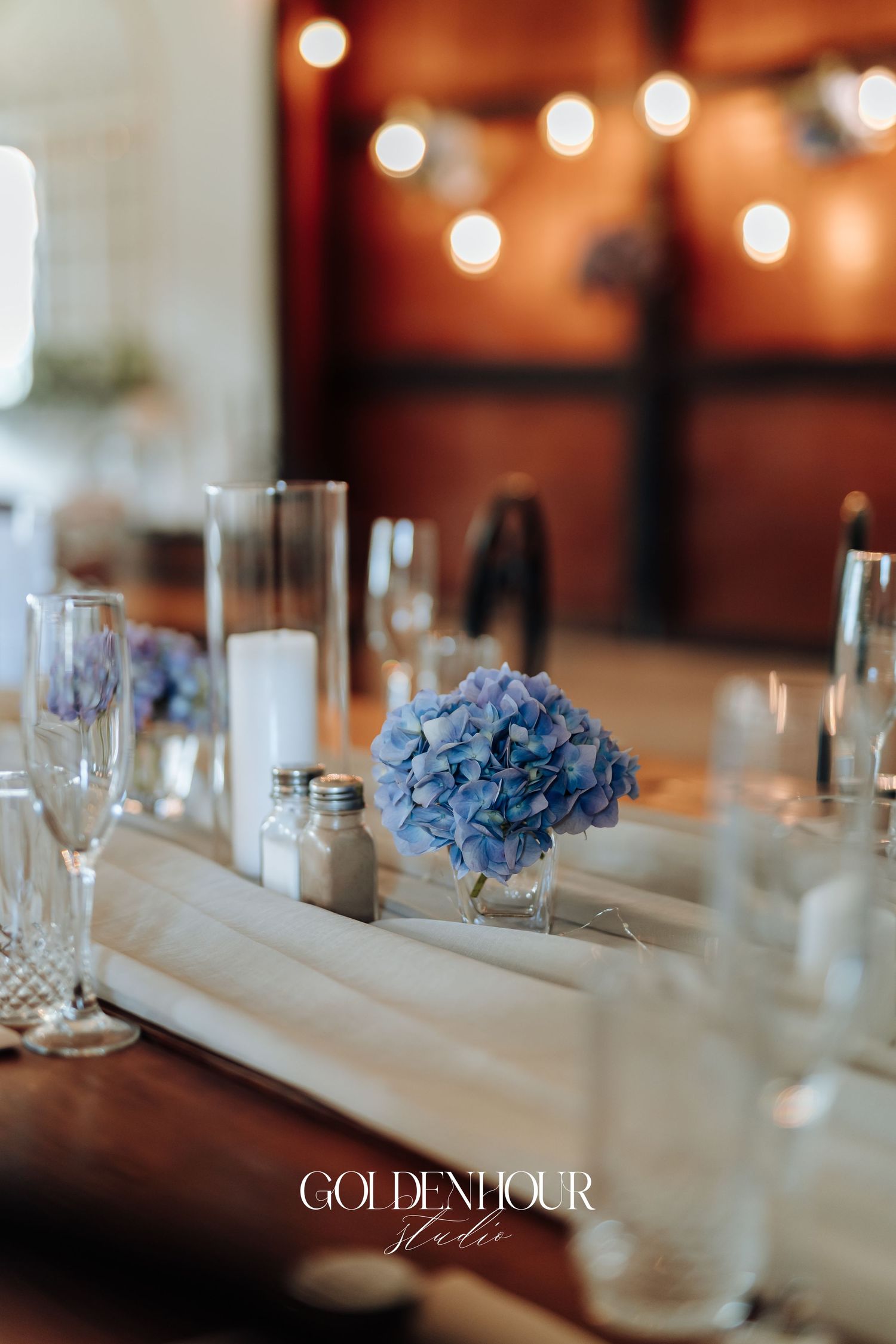 Elegant table setting with blue hydrangea centerpieces and glowing string lights in the background.