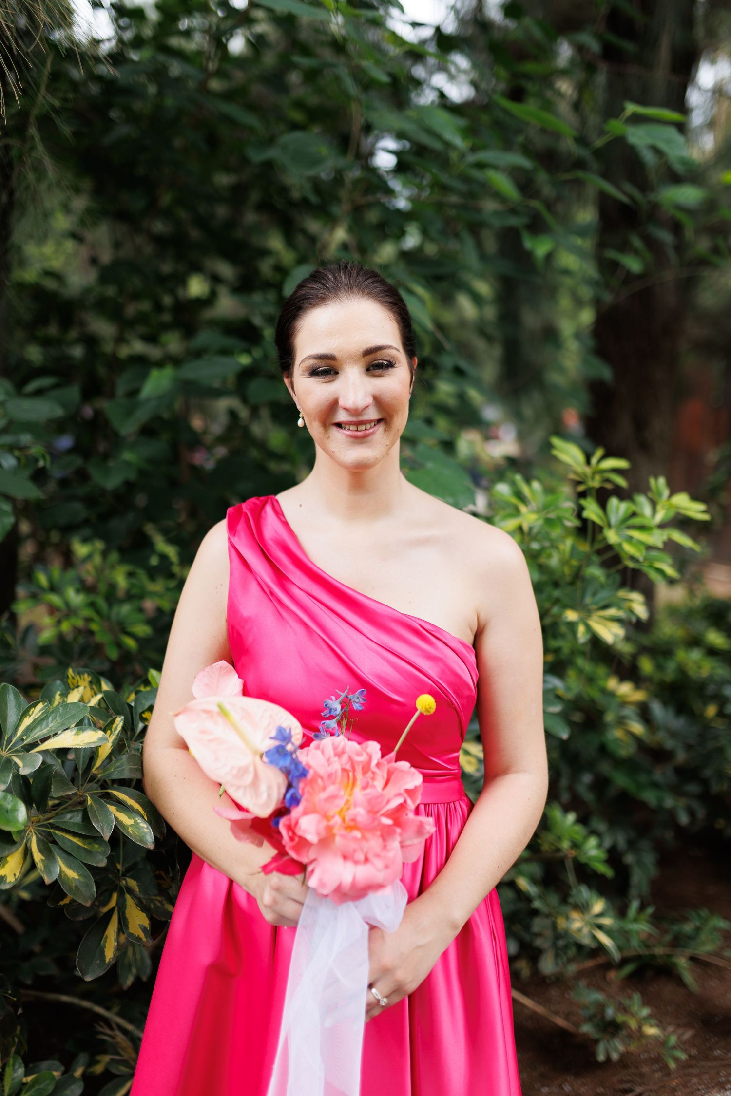 A person in a bright pink one-shoulder dress holds a colorful bouquet against a green garden backdrop.