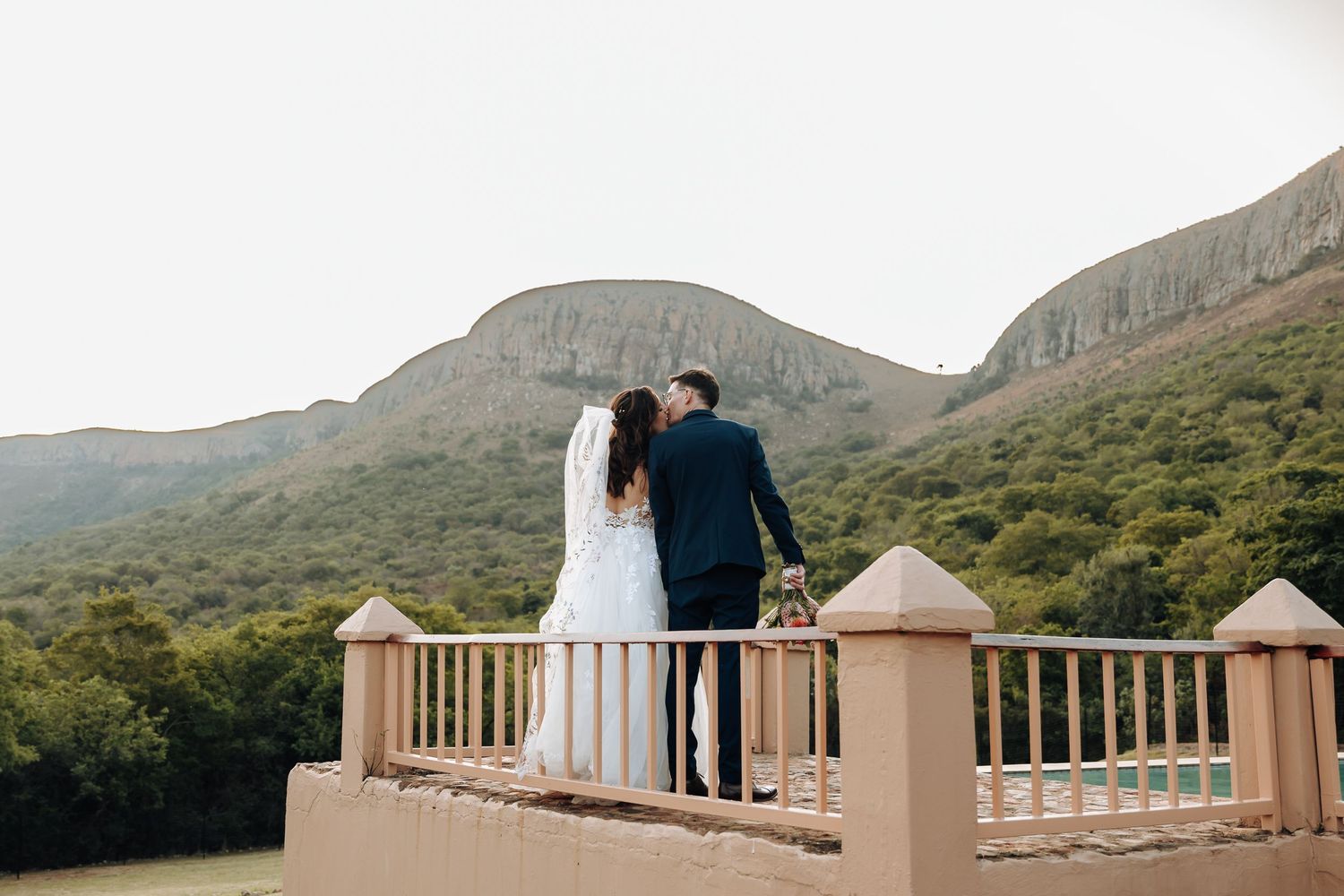 A couple stands at a scenic mountain overlook during their wedding day, with rolling hills in the background.