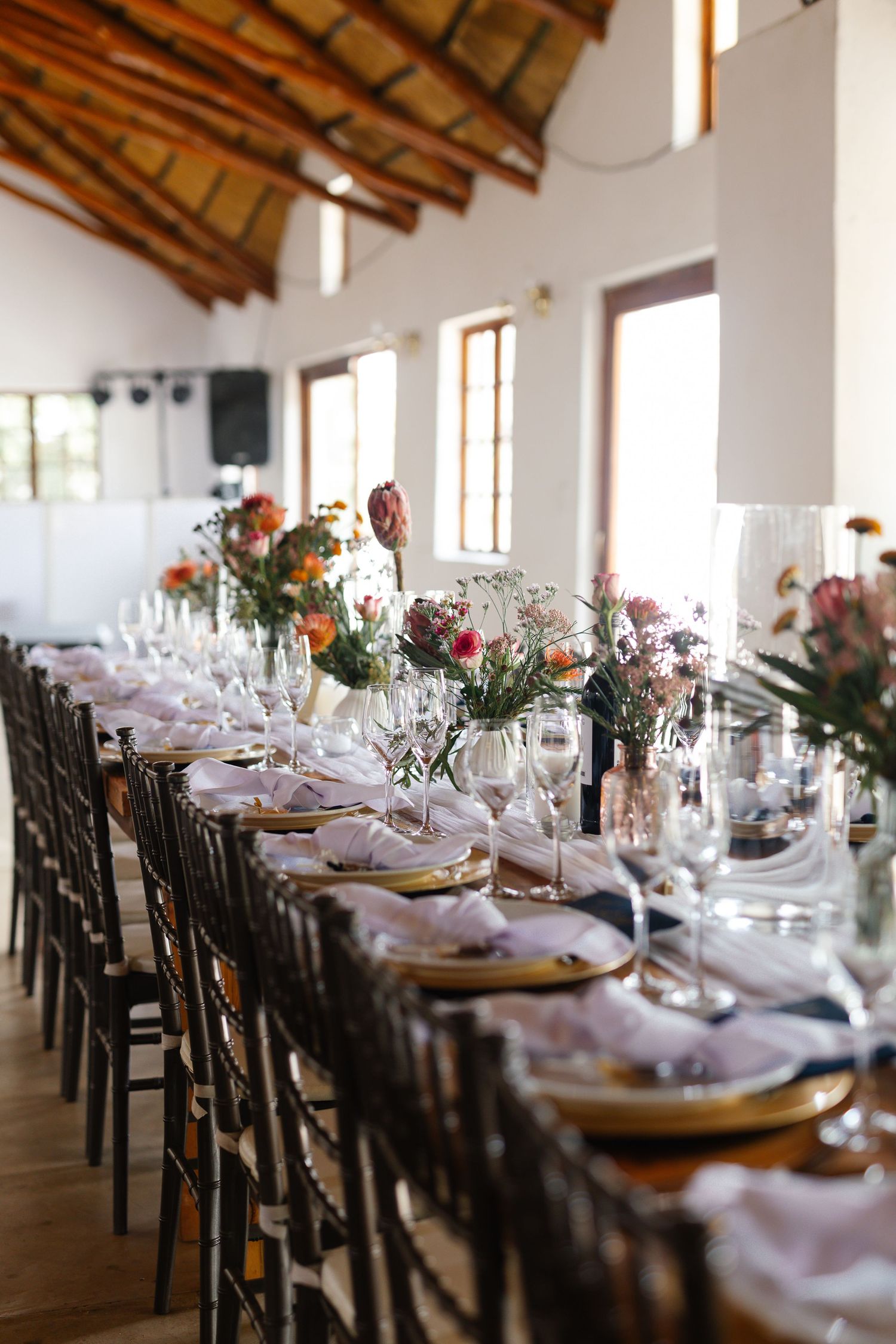 Elegant wedding reception tables set with gold chargers and floral centerpieces in a rustic venue with exposed wooden beams.