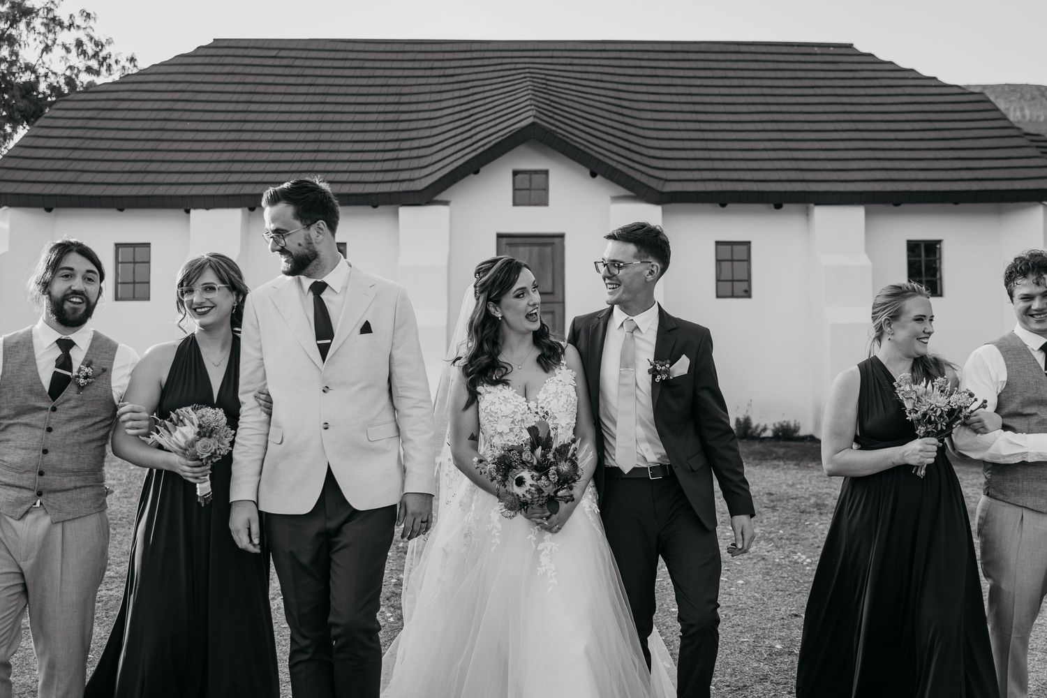 Black and white wedding party photo with bridesmaids and groomsmen standing in a line outside a house with tiled roof.