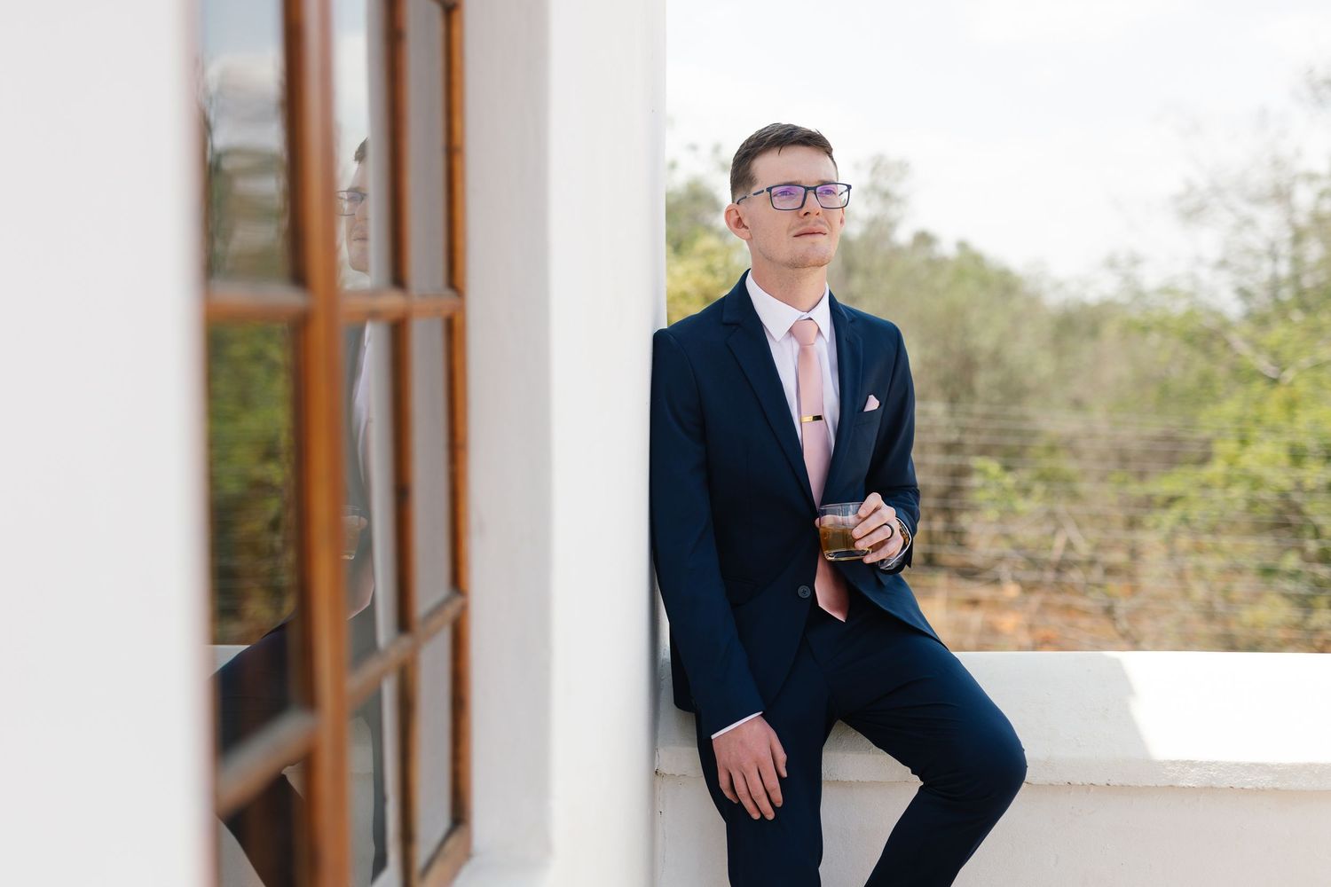 A person in a navy suit and pink tie stands confidently on a porch with white columns in natural daylight.