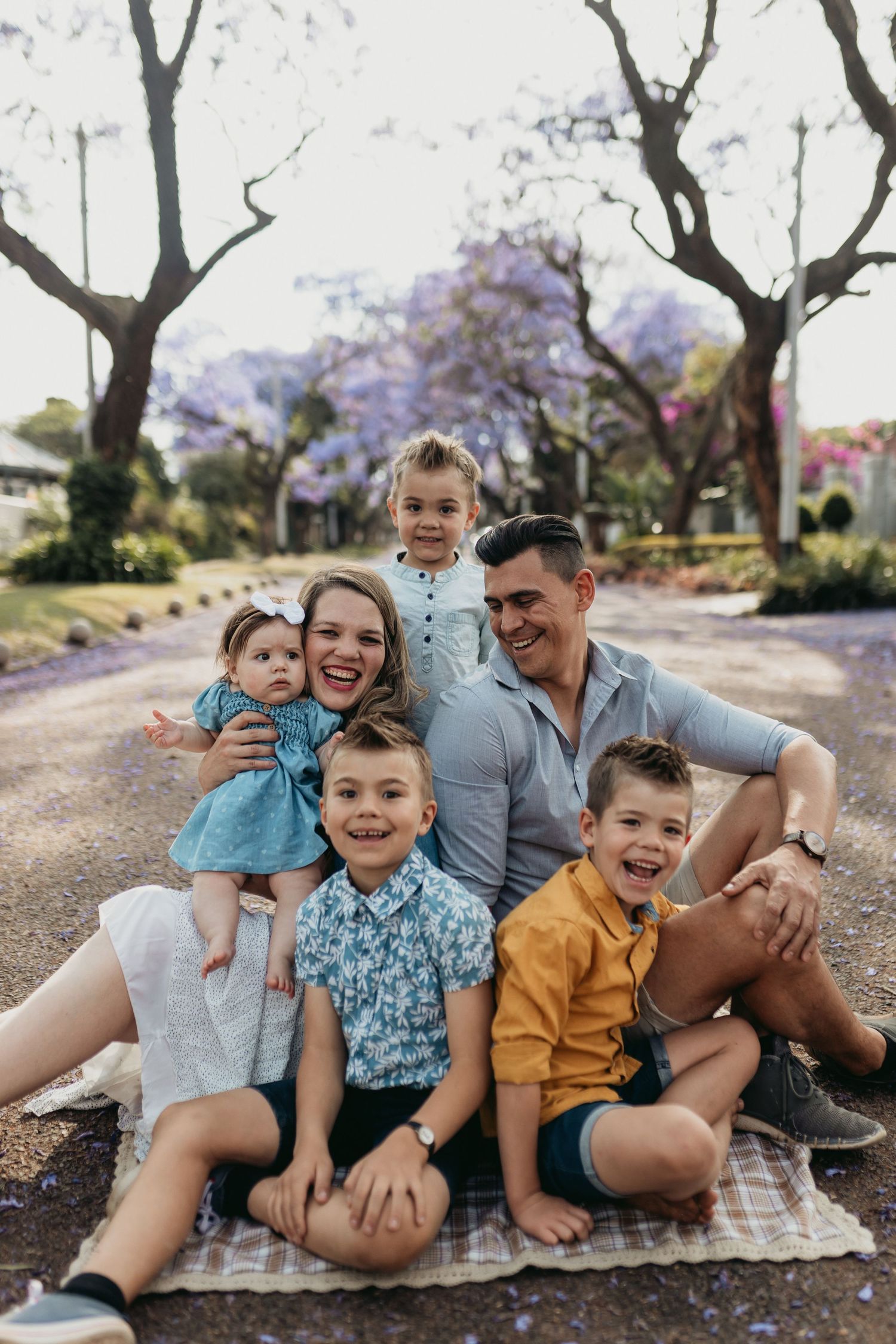 A cheerful family group lying on a cobblestone pathway surrounded by blooming jacaranda trees in the background.