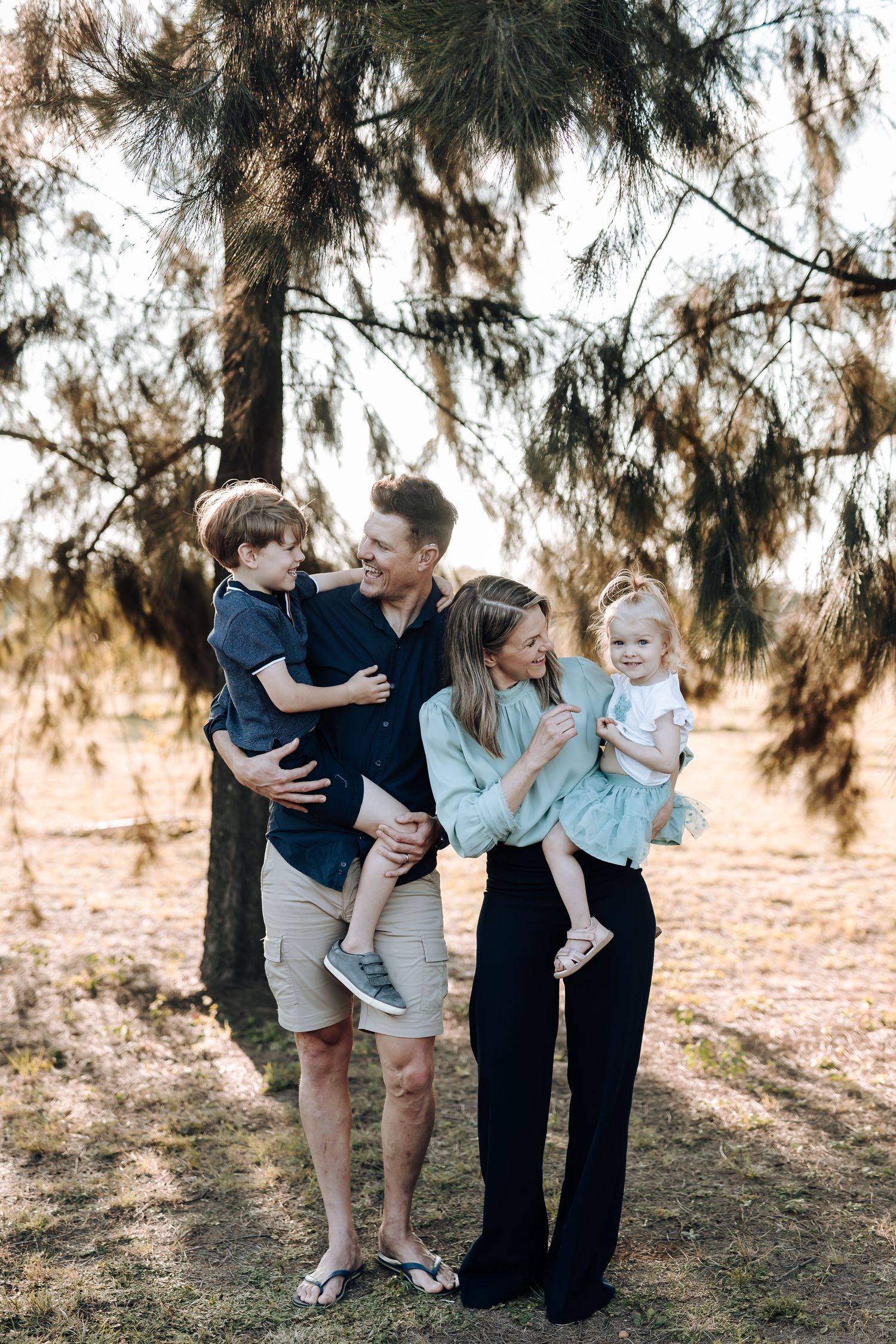 Family shares tender moments together while posing outdoors under evergreen trees in warm sunlight.