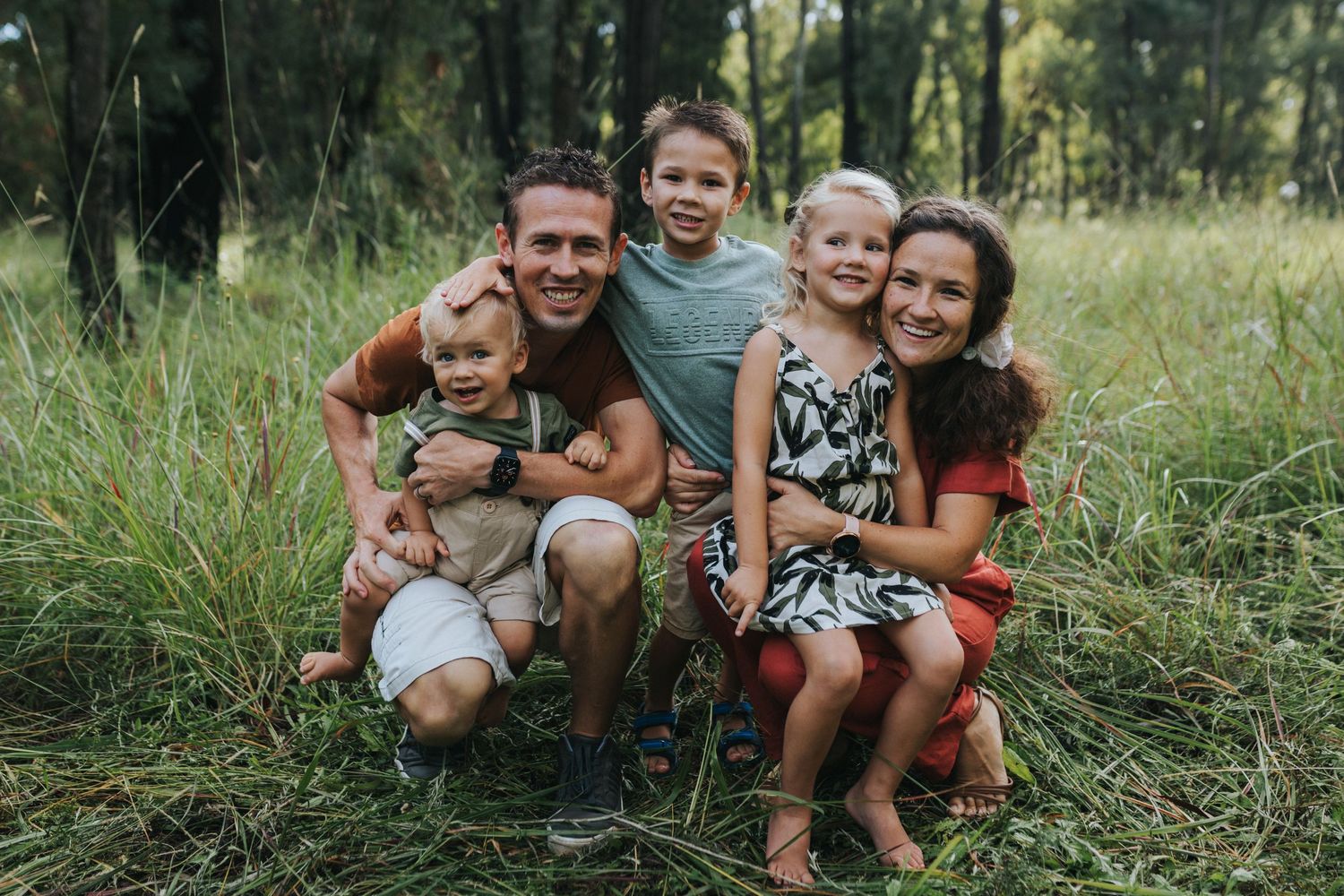 A smiling group of people sit together in a grassy meadow during golden hour lighting for a casual outdoor photo.