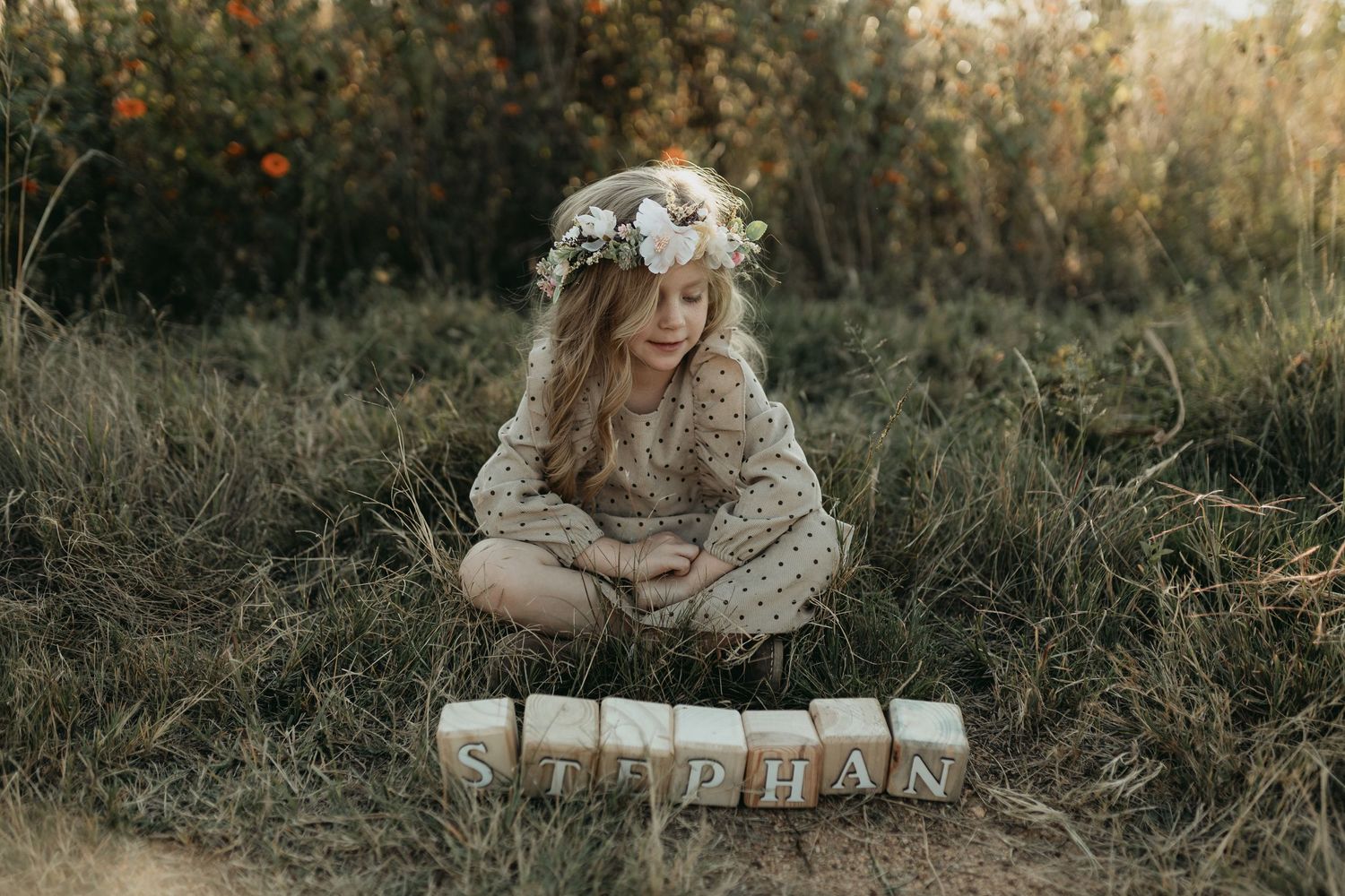 Child in floral crown and vintage dress sits beside wooden blocks on grass at sunset.