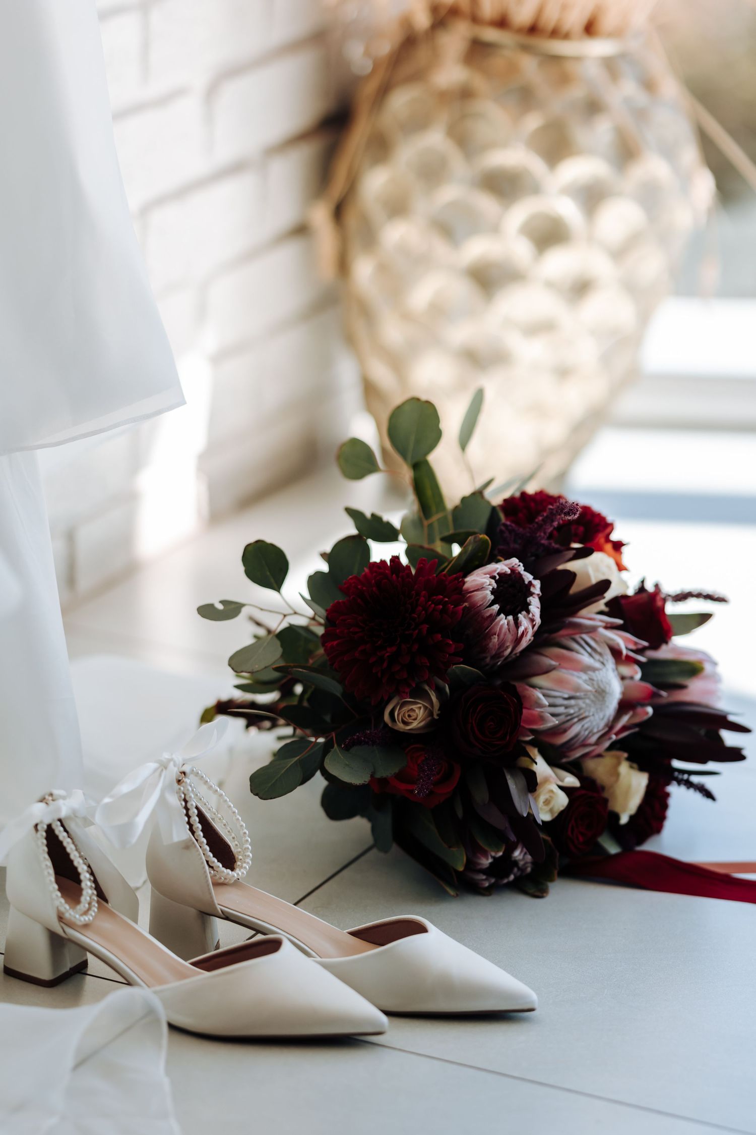 White bridal heels with pearl details beside a dark burgundy wedding bouquet with protea and eucalyptus on white floor.