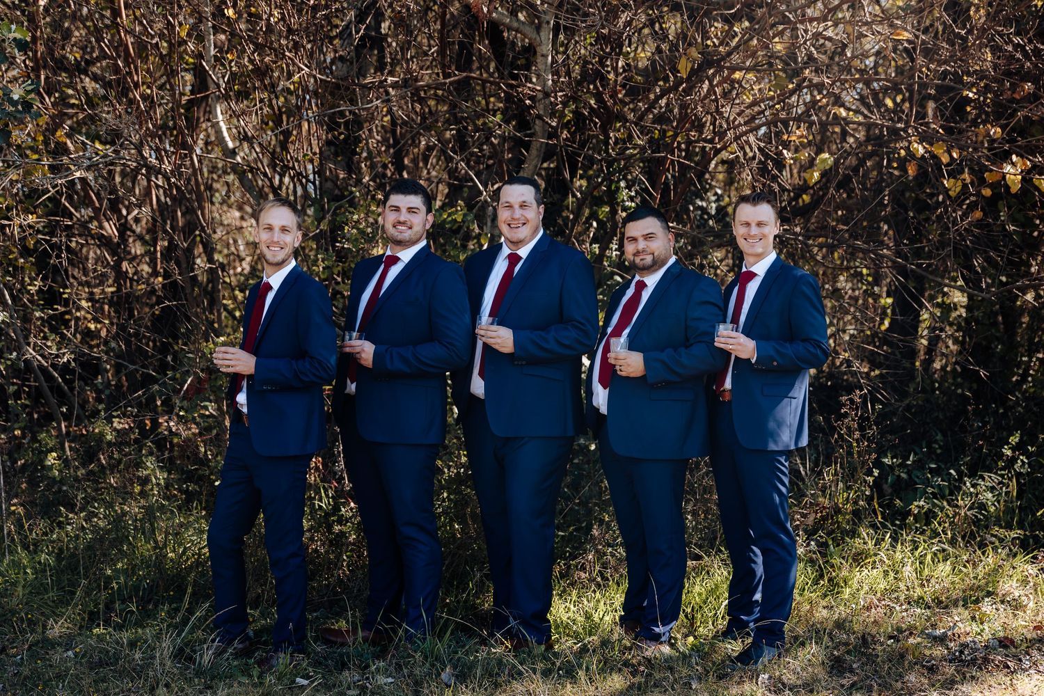 Five groomsmen in navy suits and red ties pose outdoors in front of autumn shrubs for a wedding photo.