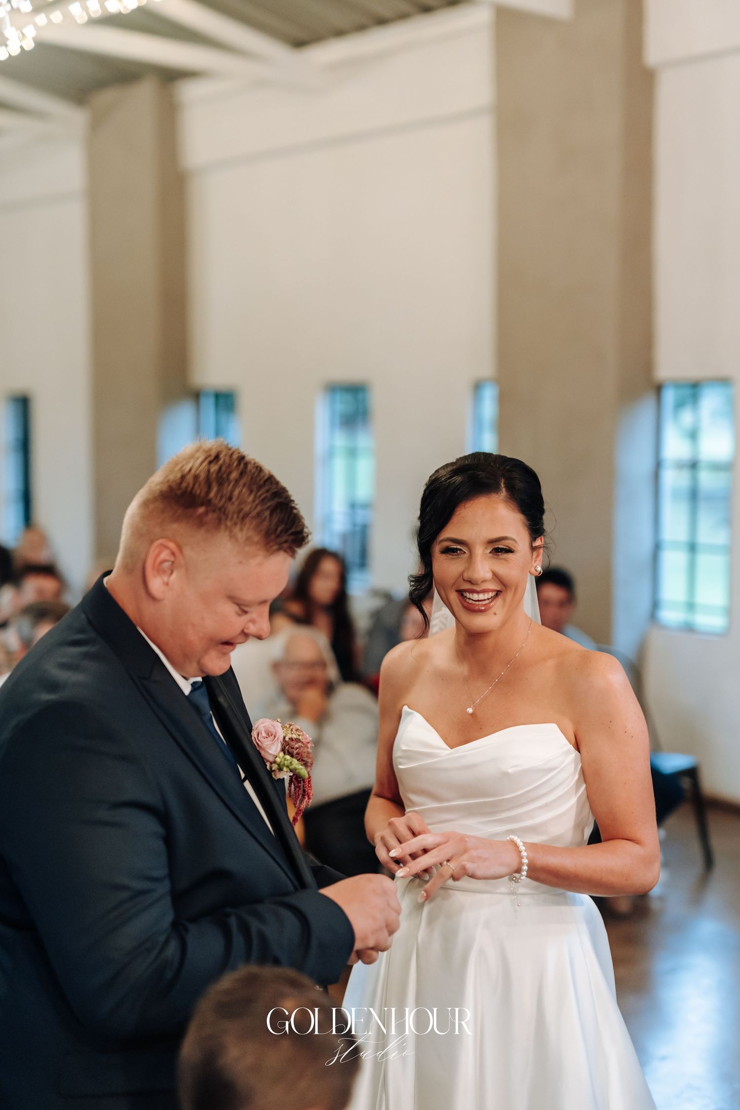 Groom placing wedding ring on smiling bride's finger during indoor wedding ceremony.