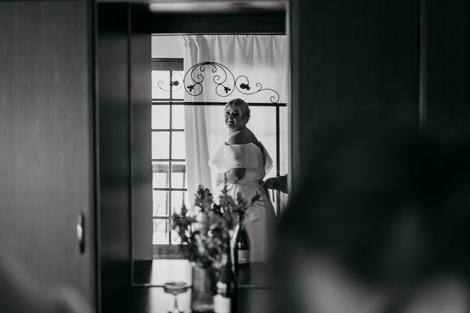 Bride in wedding dress standing by window in black and white photo, elegant bridal preparation moment.
