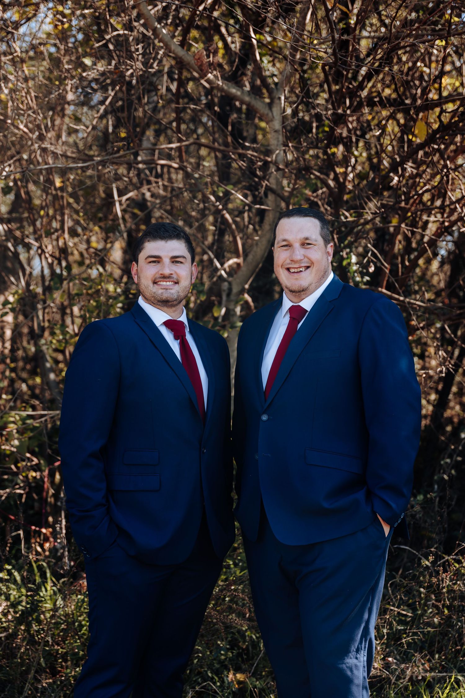 Two men in navy blue suits with red ties smiling outdoors against autumn foliage backdrop.