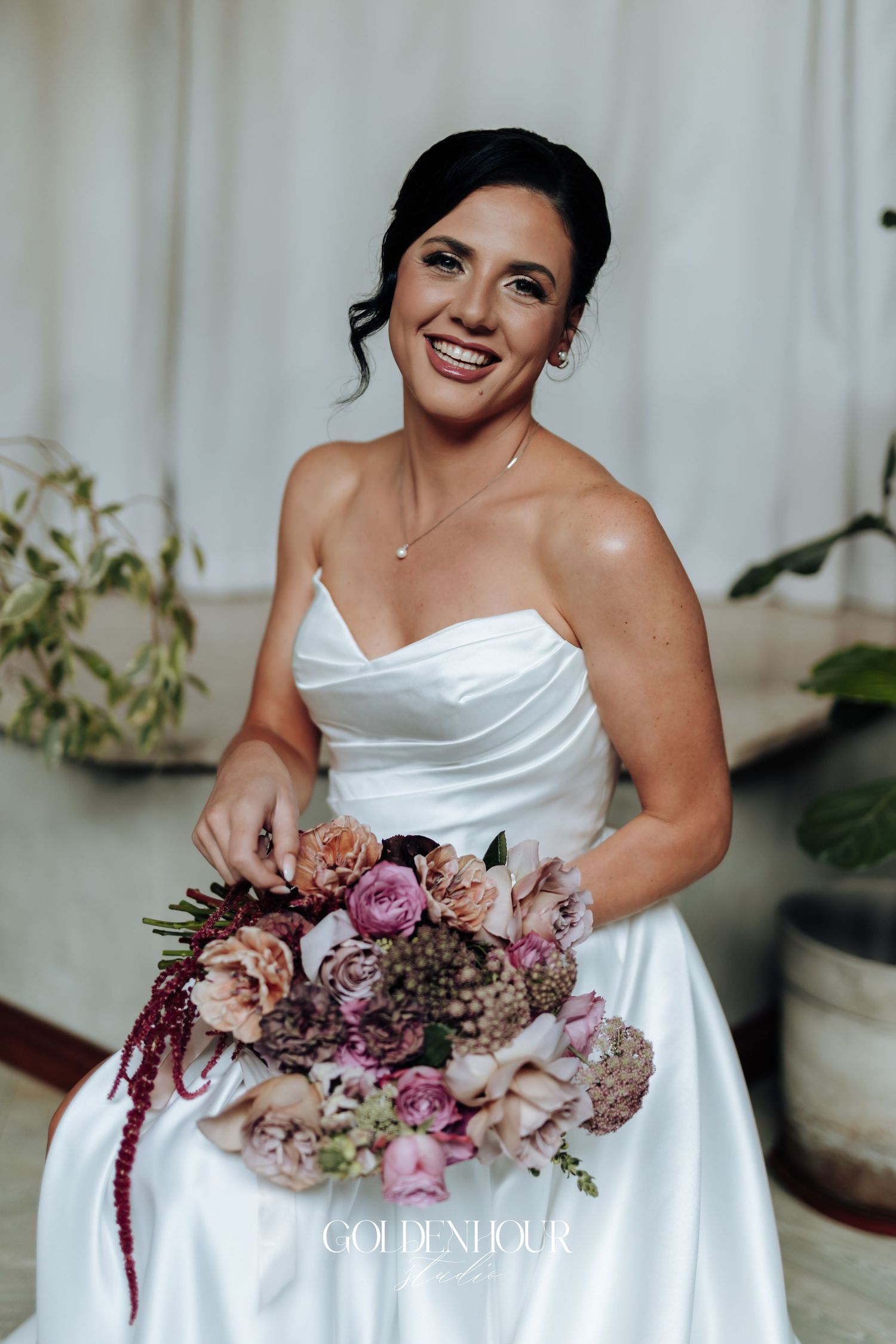 Radiant bride in strapless white satin gown holding lush mauve and burgundy wedding bouquet and smiling.