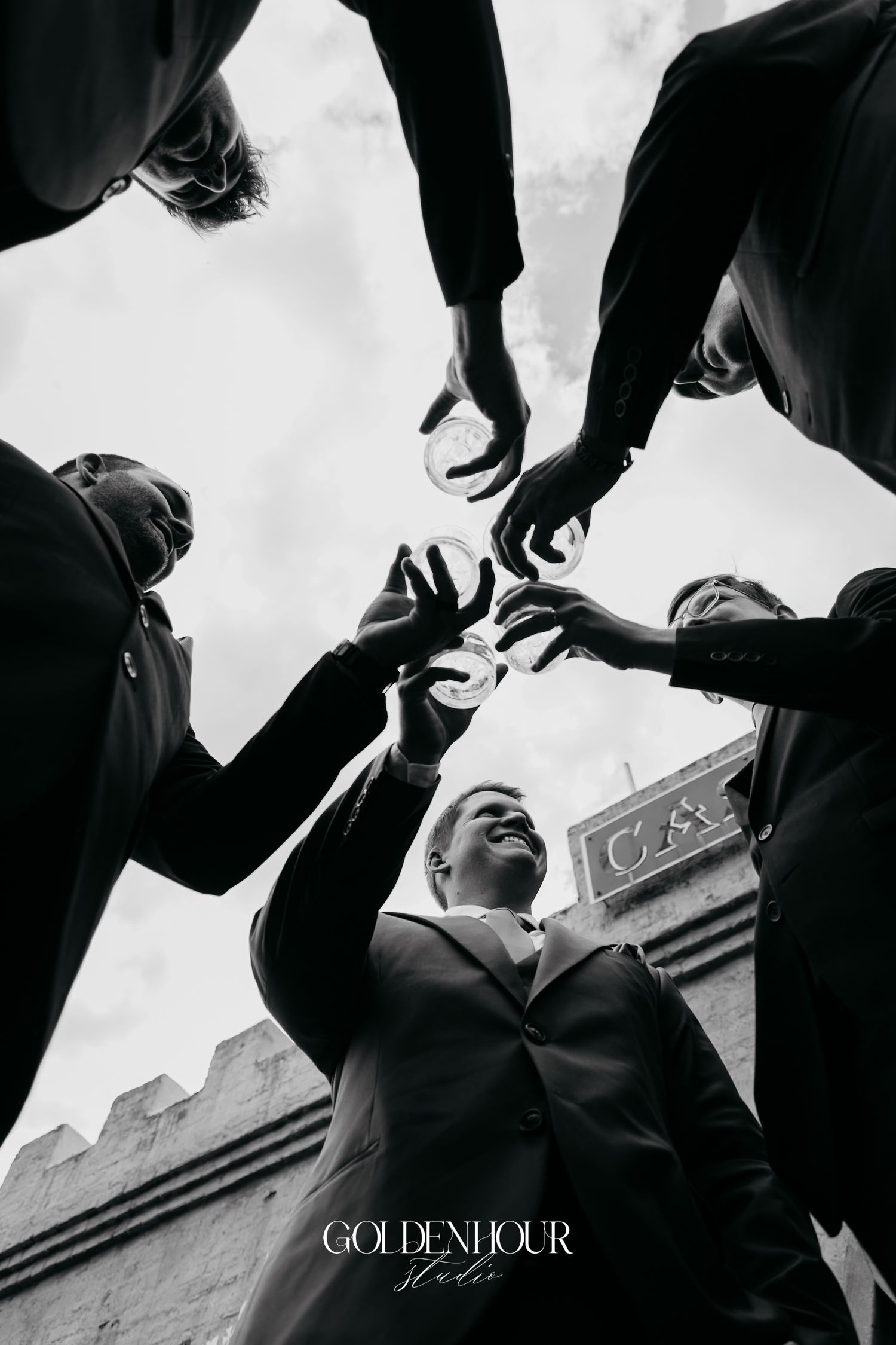 Creative low-angle black and white shot of groomsmen reaching toward the groom holding wedding rings outdoors.