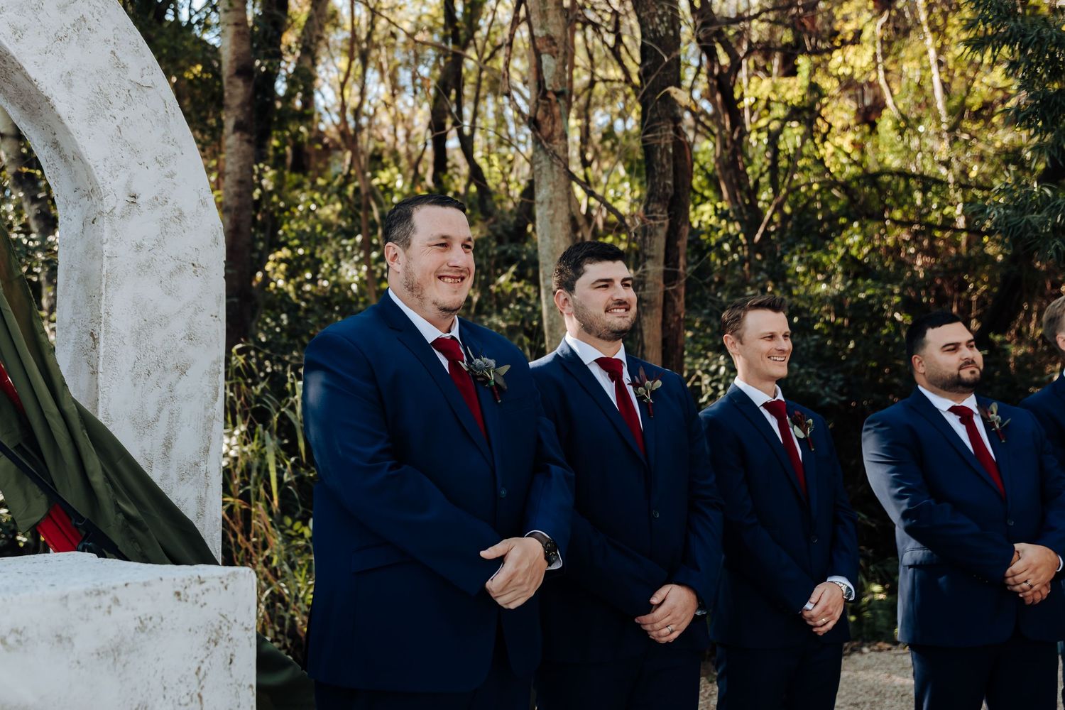 Four groomsmen in navy suits with red boutonnieres stand smiling at an outdoor wedding ceremony surrounded by trees.