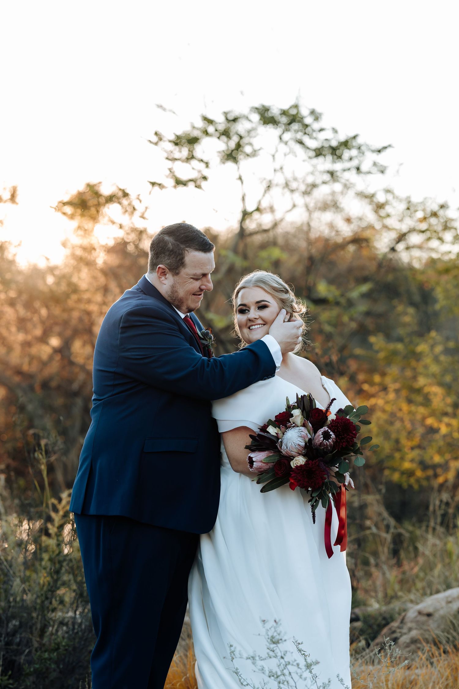 Bride and groom embrace outdoors at golden hour, she holds a burgundy bouquet, he wears a navy suit, autumn trees behind them.
