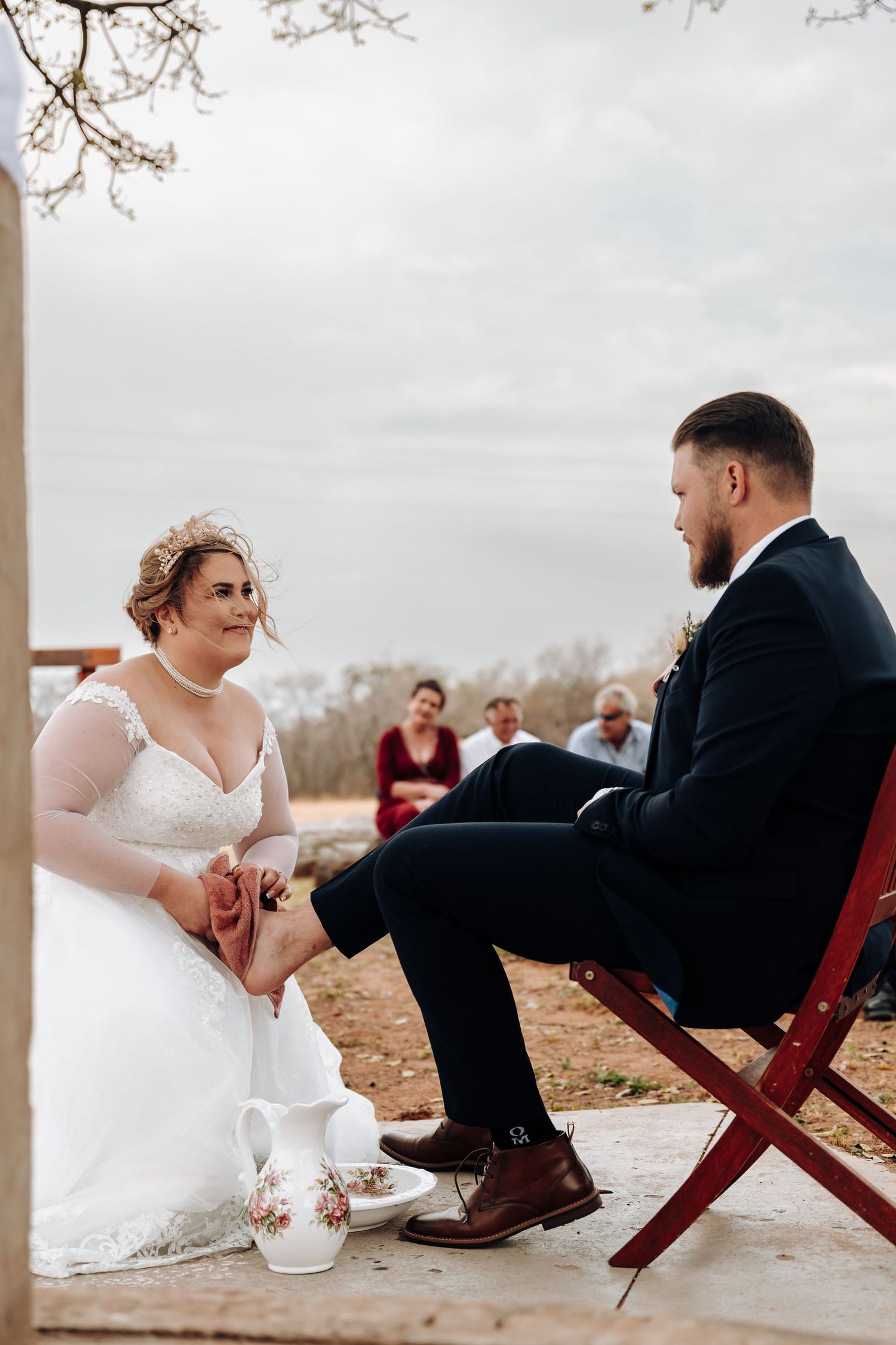 A romantic wedding scene showing the bride and groom sitting on red chairs during their rustic outdoor ceremony.