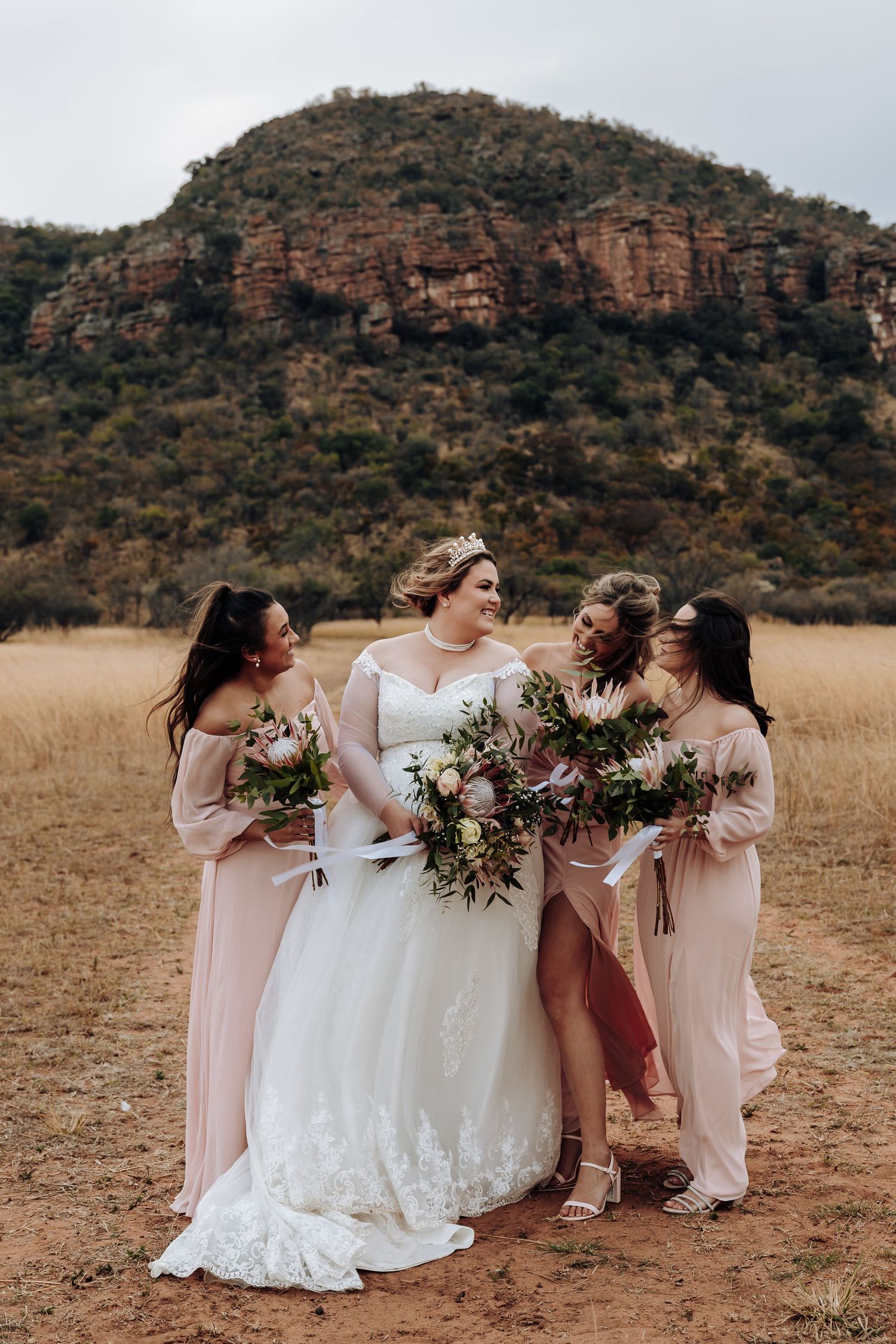 Wedding party wearing blush pink dresses and white gown hold bouquets against dramatic rocky mountain backdrop.