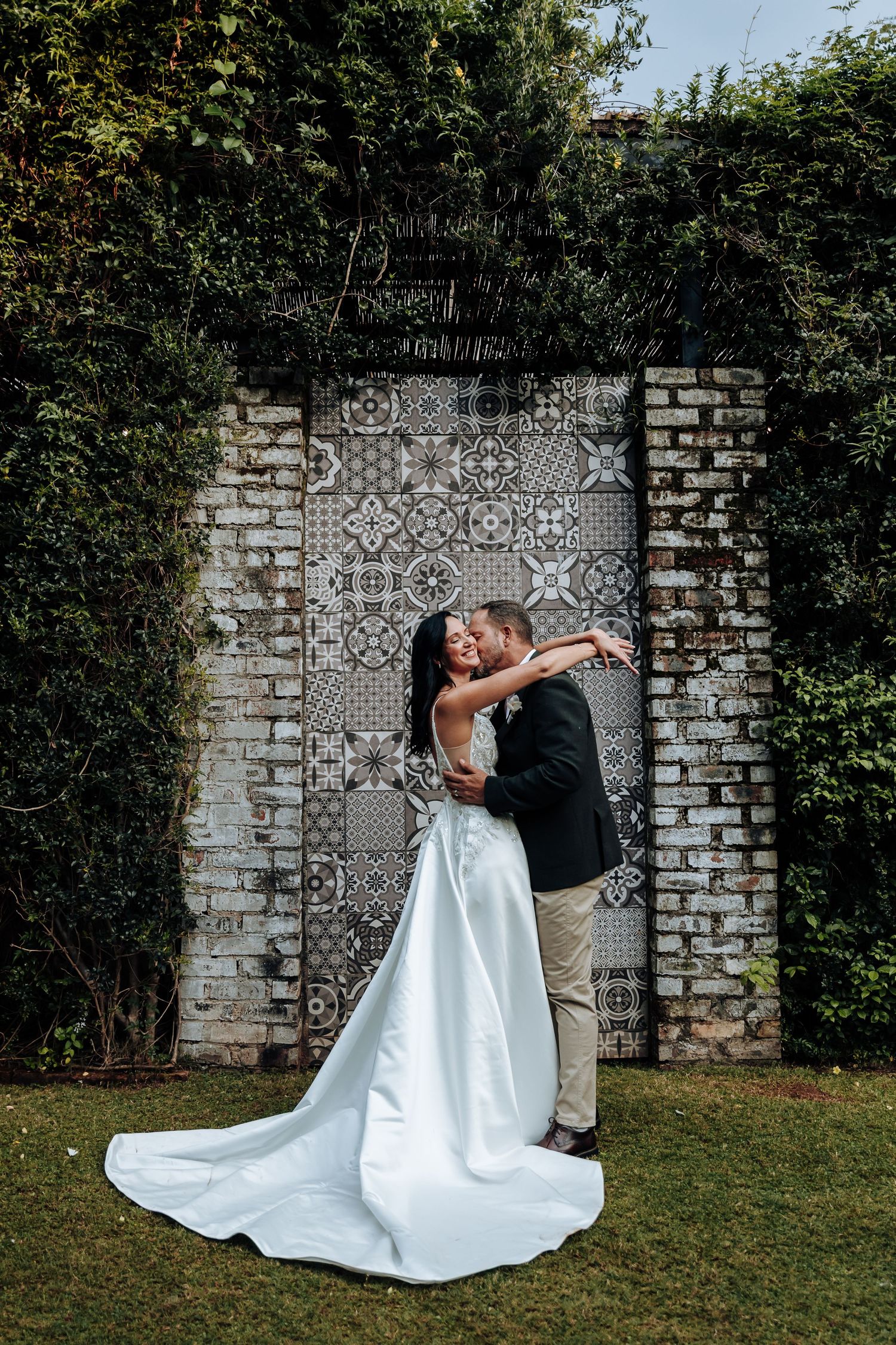 Bride and groom sharing a romantic kiss in front of a decorative tiled wall surrounded by lush greenery.