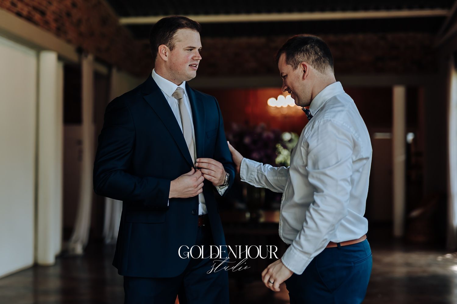 Two men in formal attire help each other prepare before a wedding ceremony indoors.