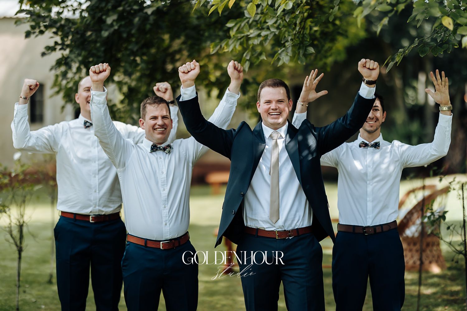 Groom and groomsmen in navy suits celebrate with raised fists outdoors at a wedding.