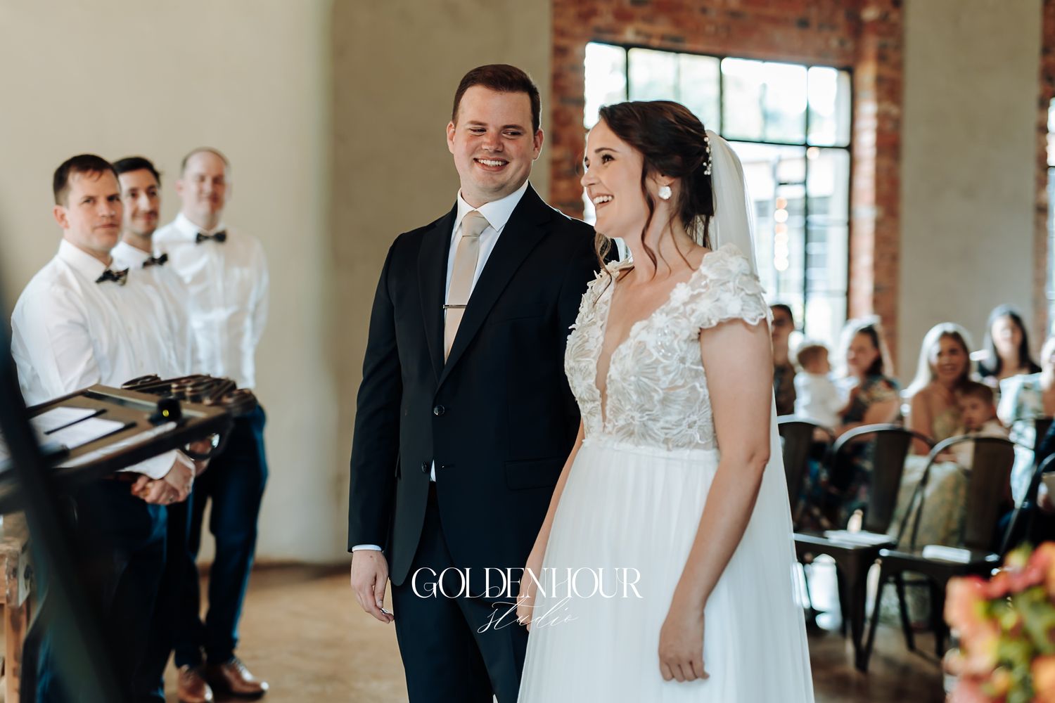 Bride and groom smiling together at the altar during indoor wedding ceremony with guests seated.