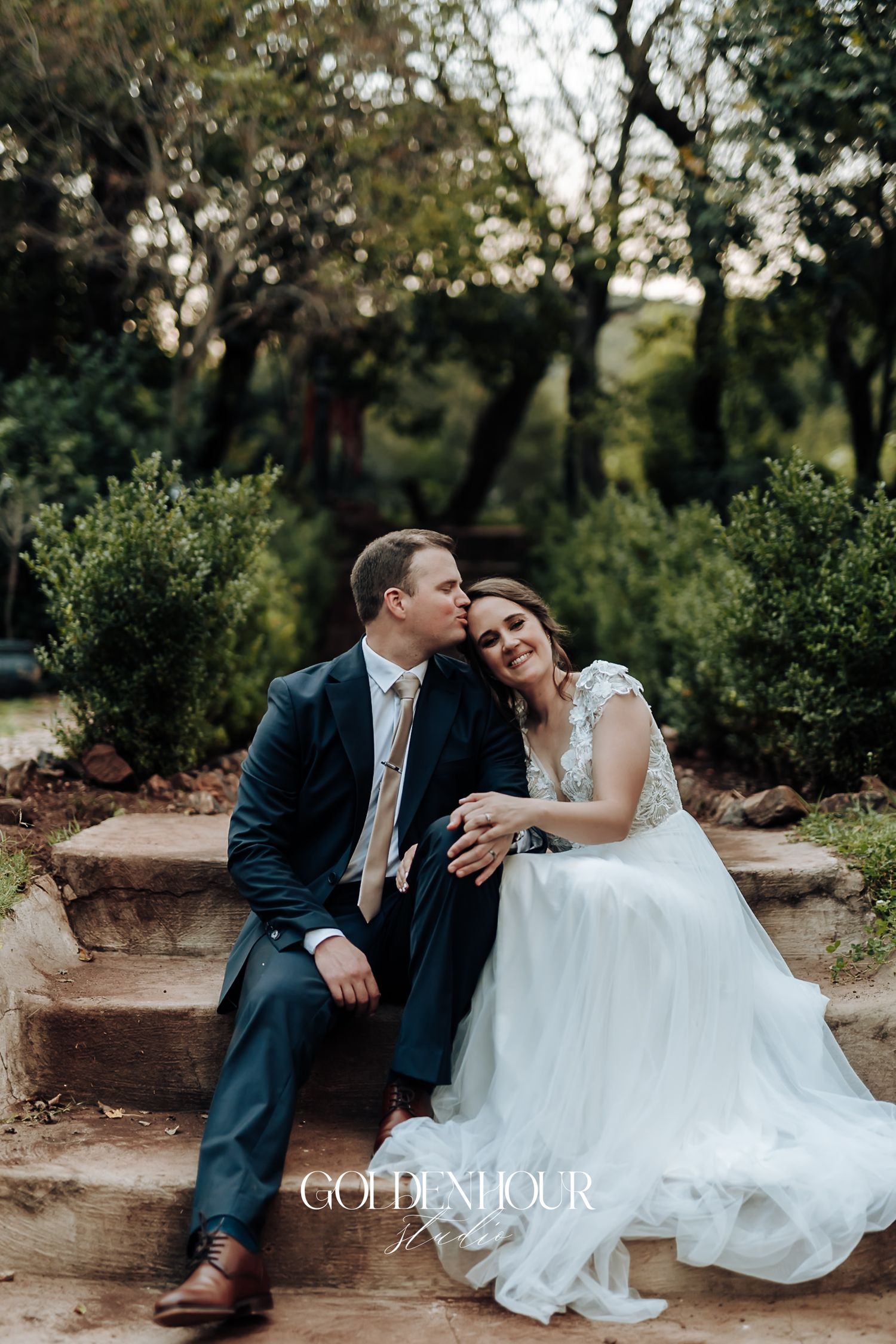 Bride and groom sit closely on stone steps, sharing a tender kiss in a lush garden setting.