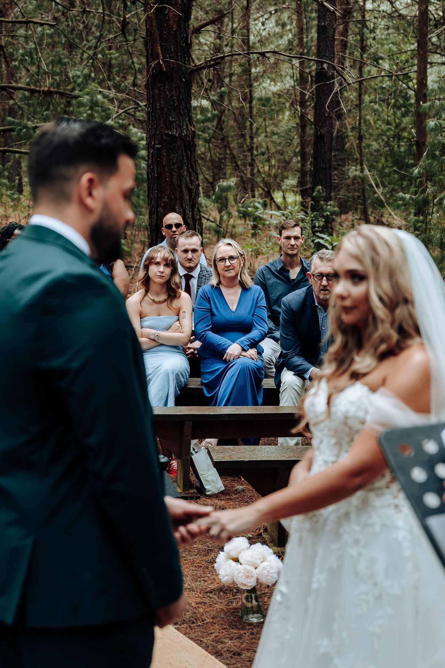 An intimate forest wedding ceremony takes place with guests seated on wooden benches among tall pine trees.