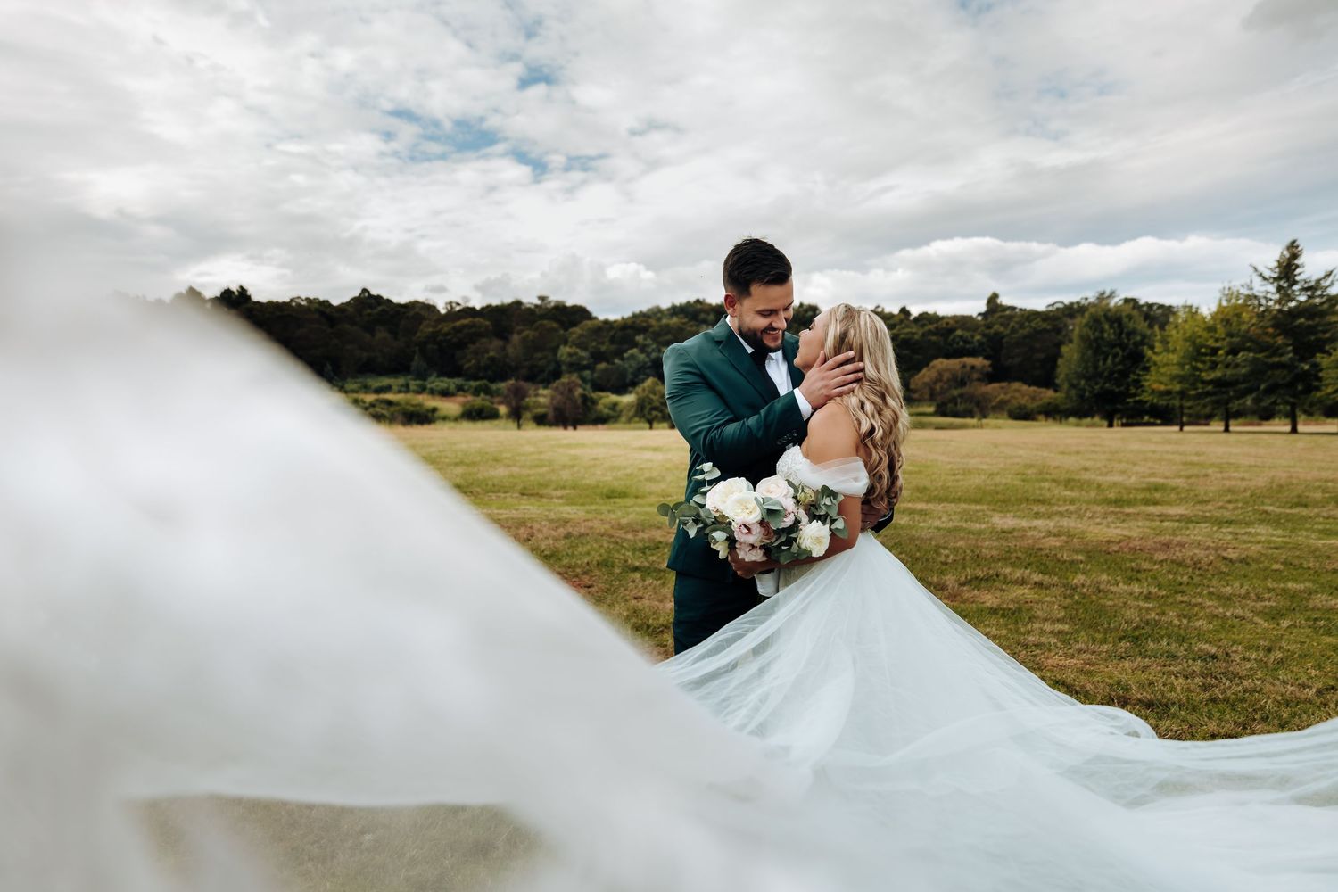 Couple in elegant wedding attire embraces in a scenic mountain meadow with dramatic cloudy skies above.