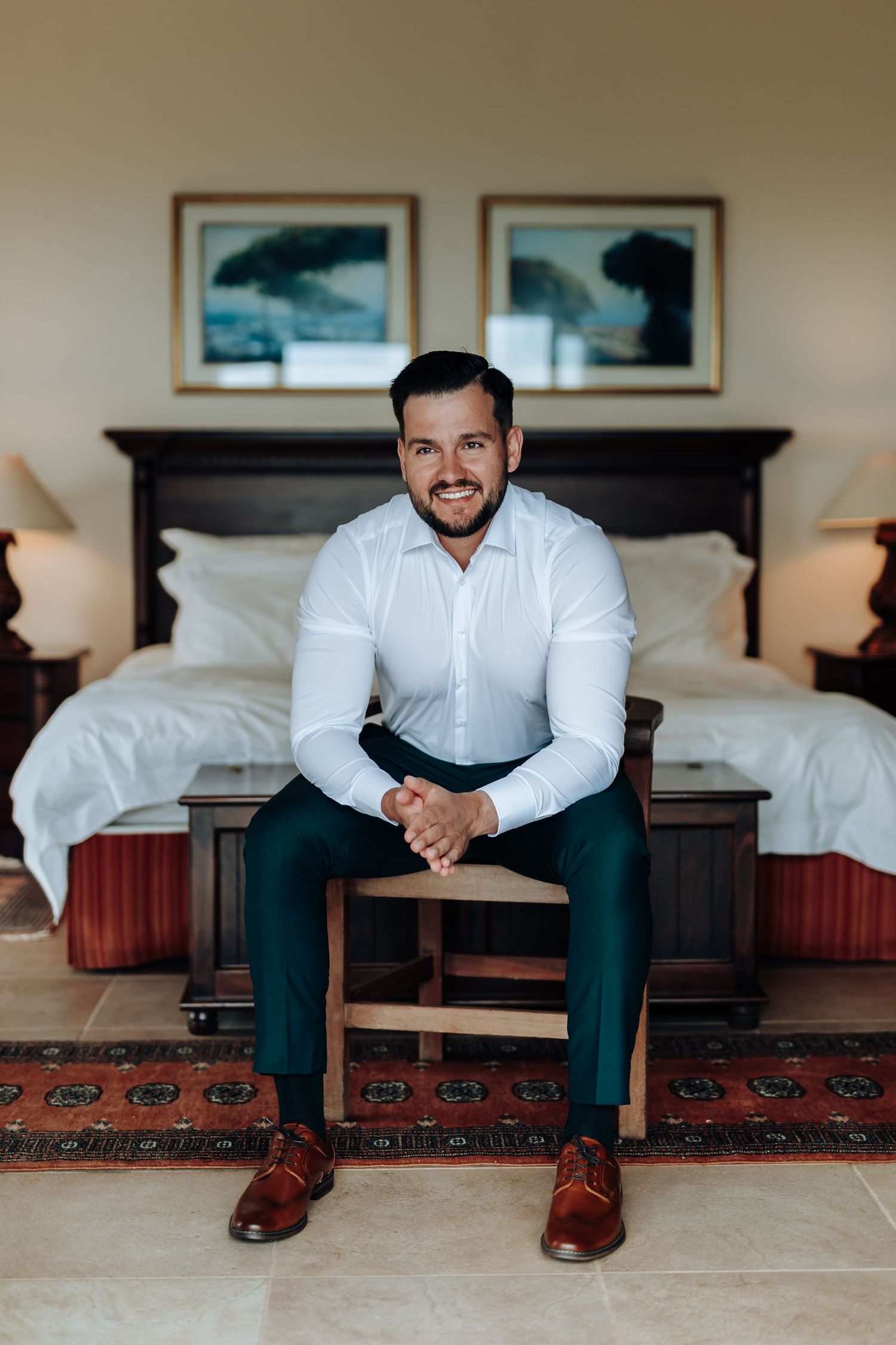 A person in a white shirt and dark pants sits on a wooden bench in a hotel room.