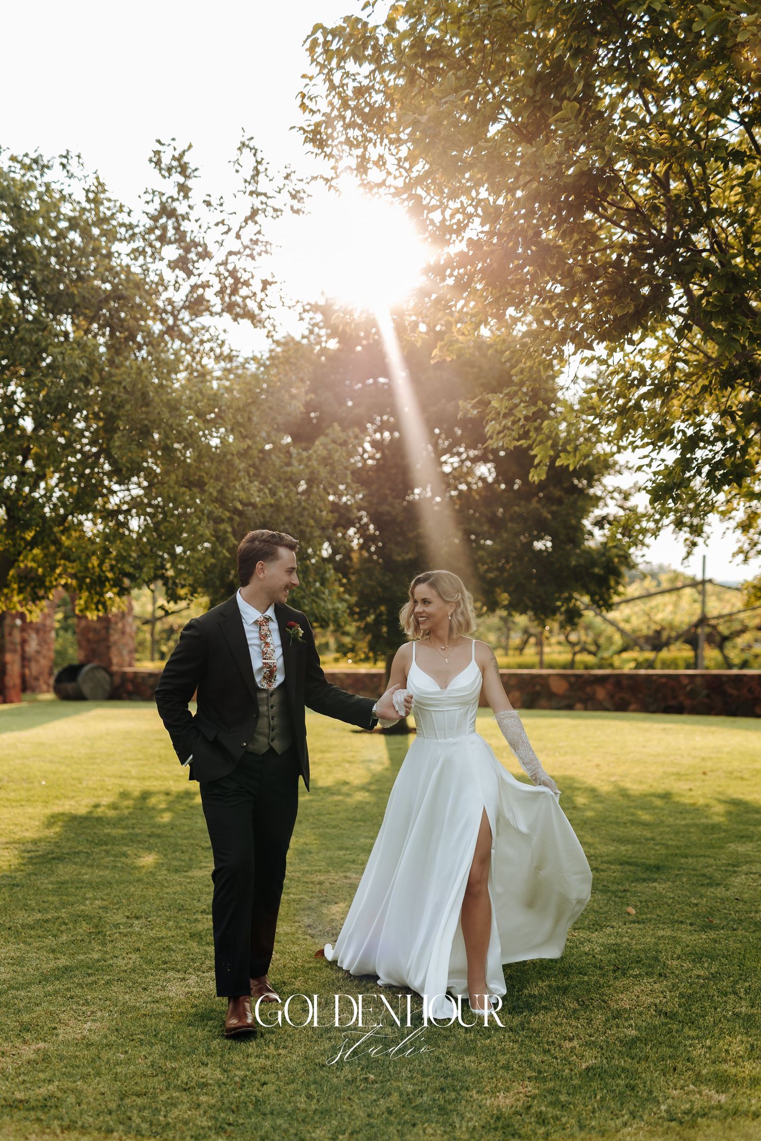 Newlyweds walk hand in hand through sunlit garden during golden hour.