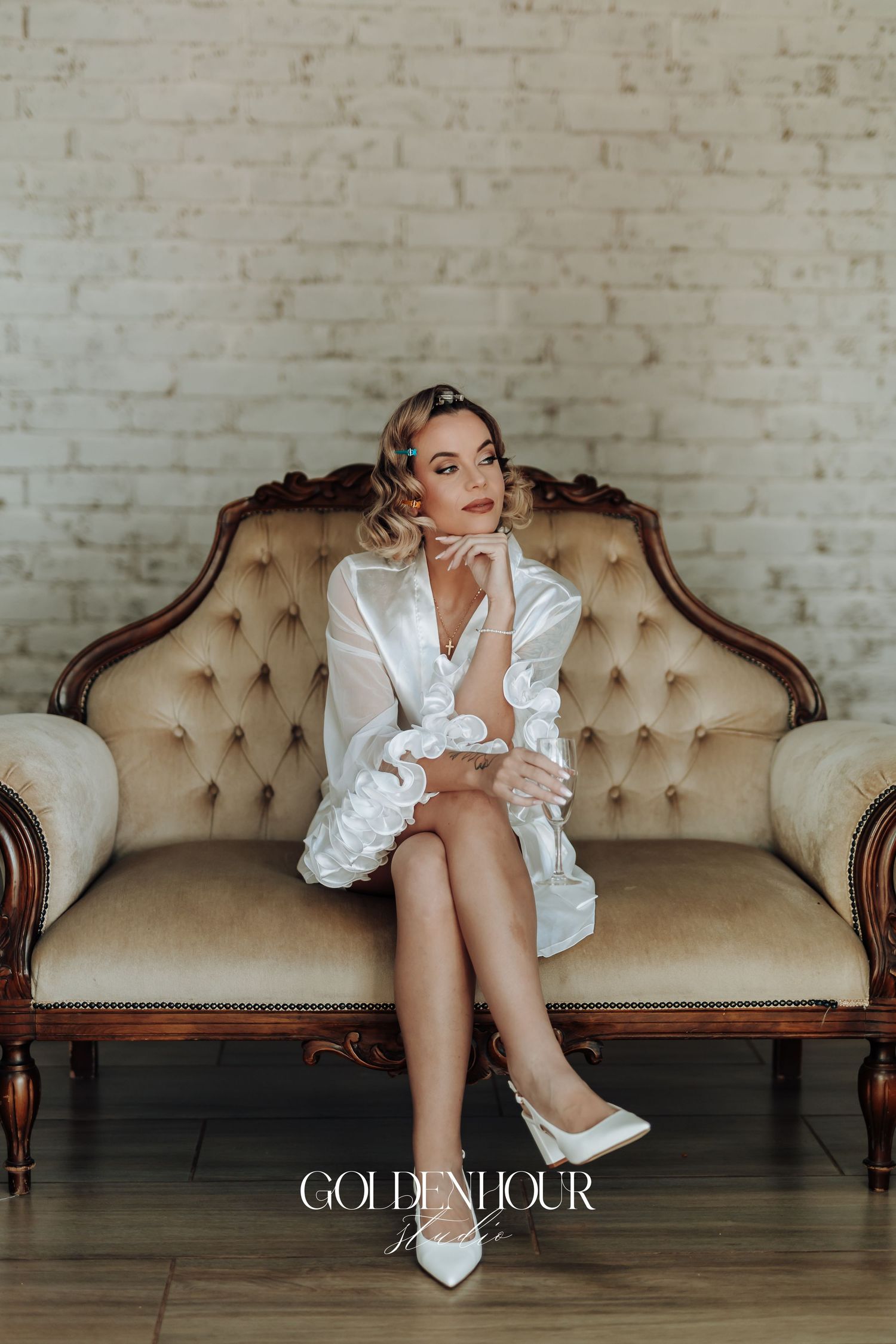 Artistic boudoir session with white silk robe on a beige tufted vintage sofa against brick wall.