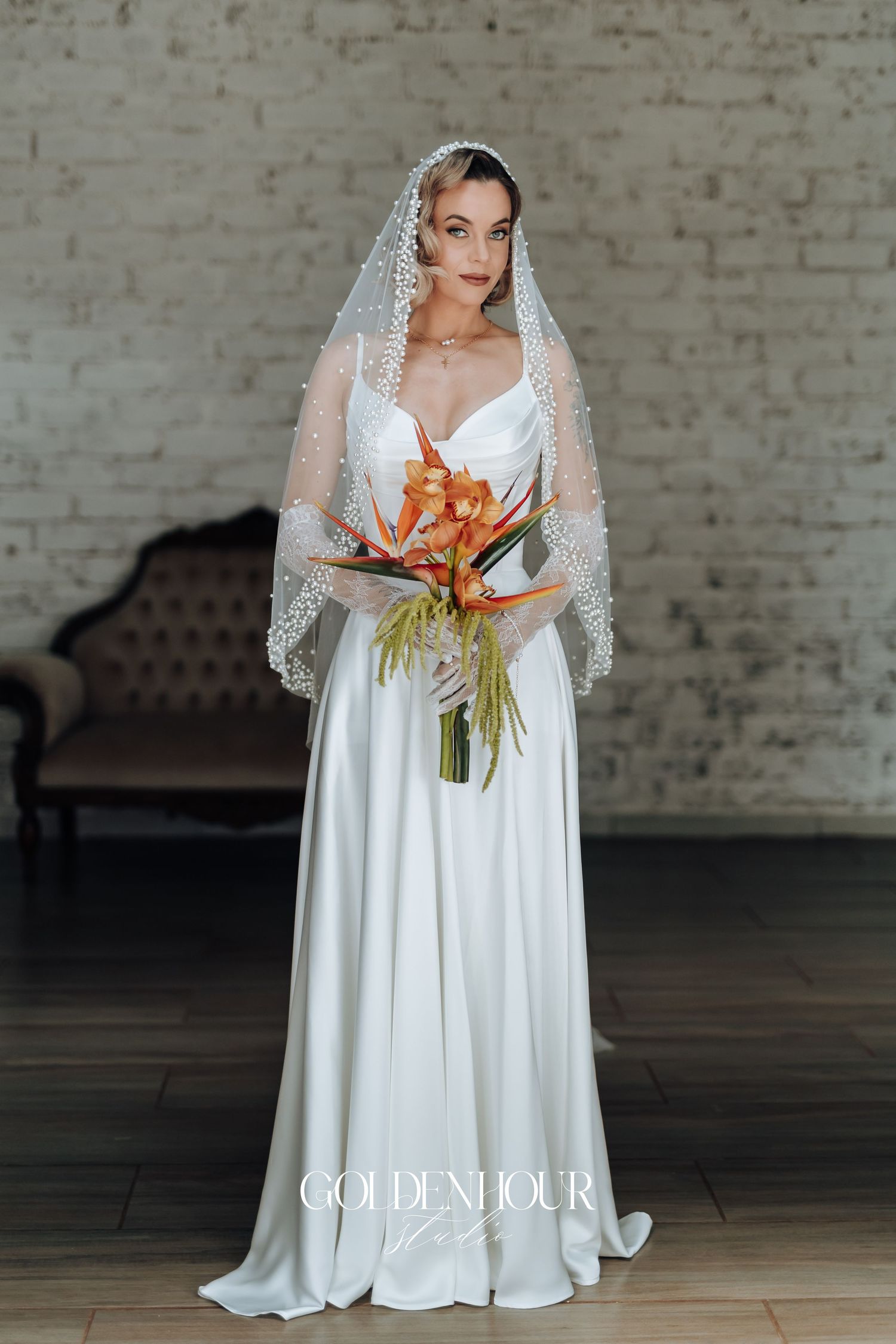 Bride in elegant white gown holds tropical bouquet against white brick wall backdrop.
