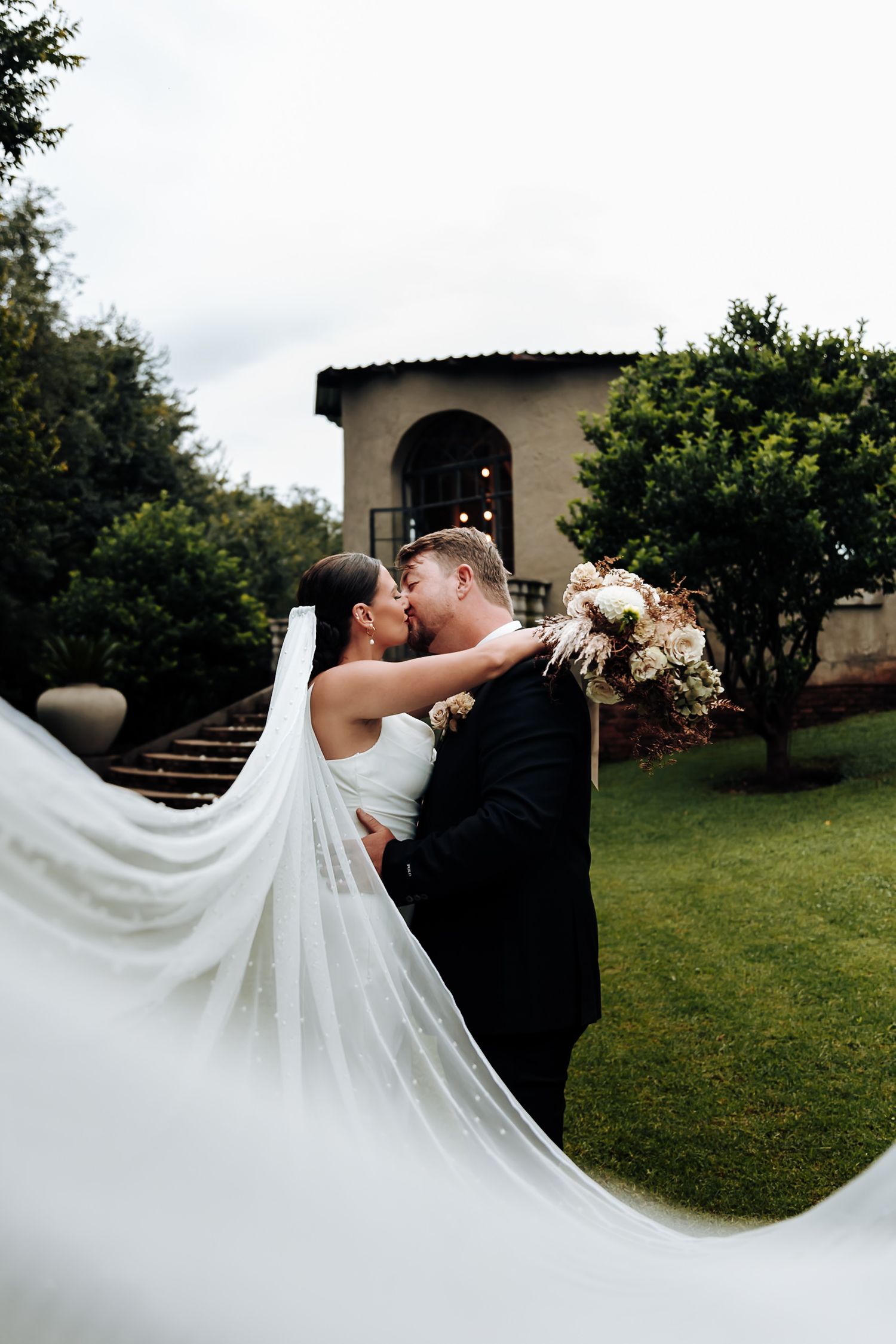 A couple embraces in a romantic kiss outdoors with a flowing white wedding dress veil dramatically sweeping behind them.