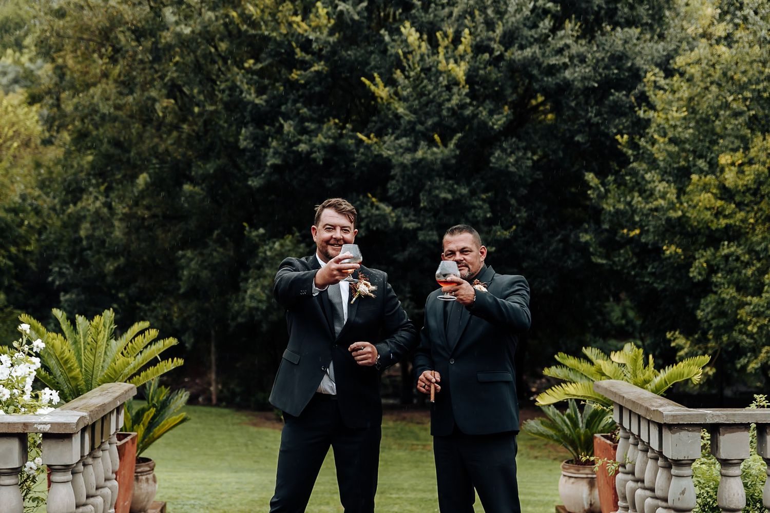 A wedding party poses in formal attire outdoors in a garden setting with greenery and stone architecture.