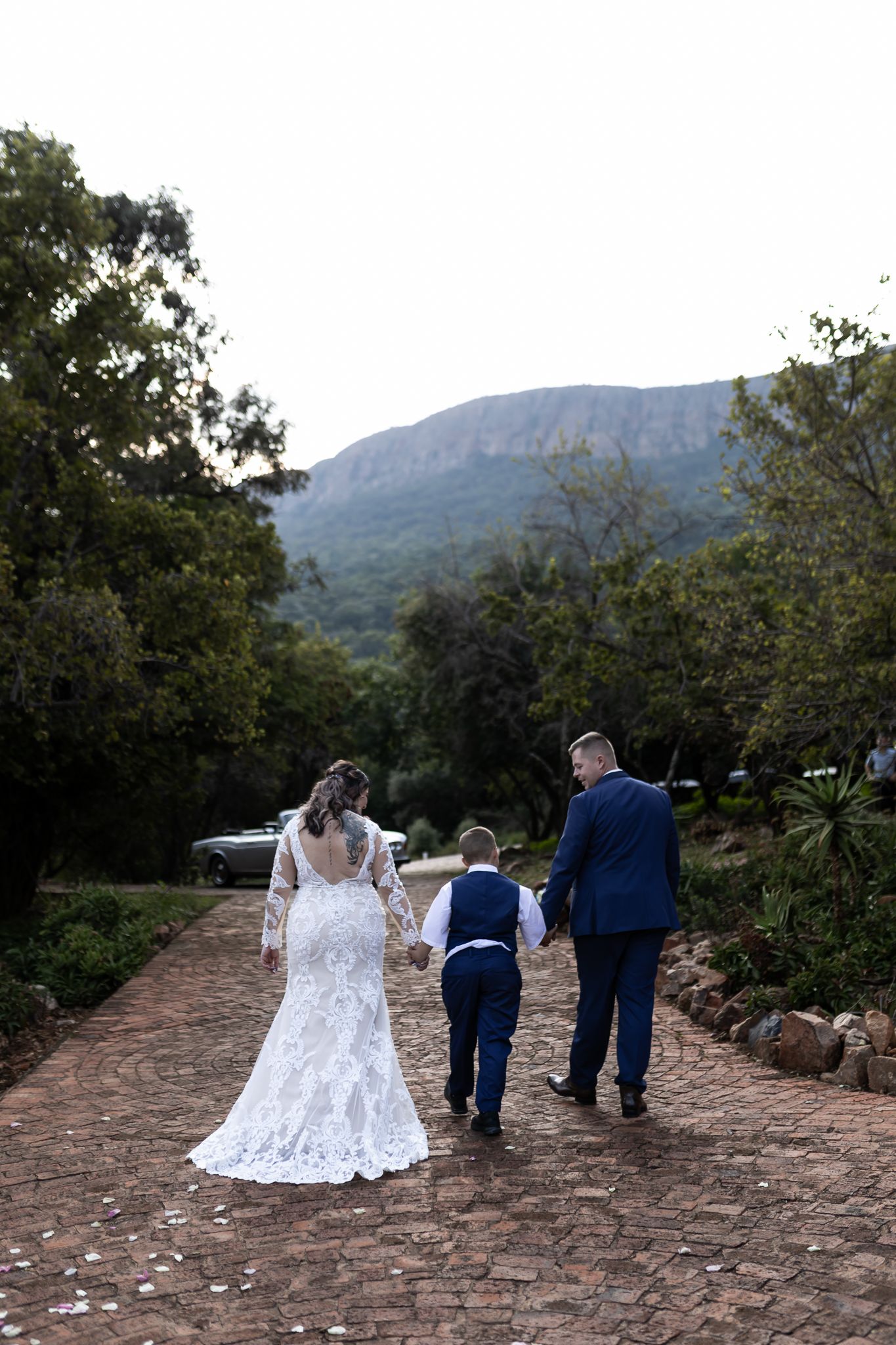 Newlyweds walk with young boy down stone path with mountain backdrop.