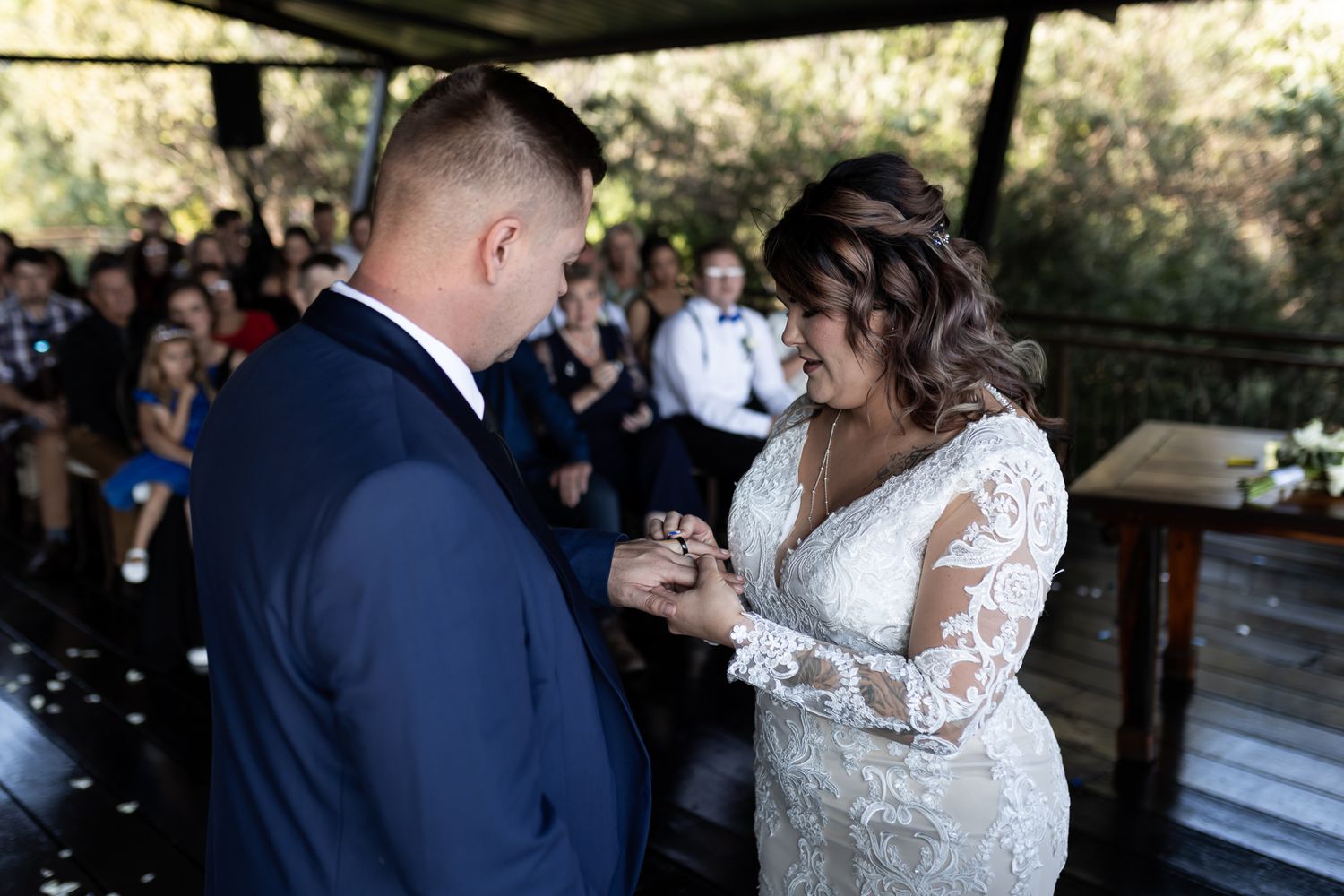 Newlyweds exchange rings during outdoor ceremony under covered venue with guests watching.