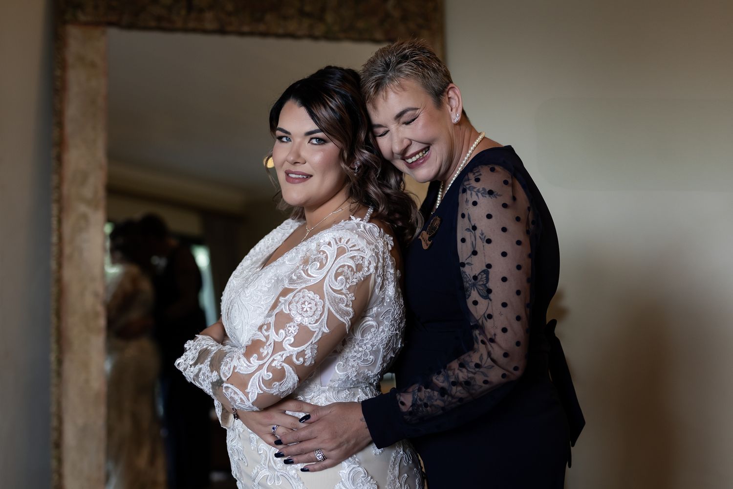 Bride in white lace wedding dress shares a joyful moment with companion wearing polka dot top during preparation.