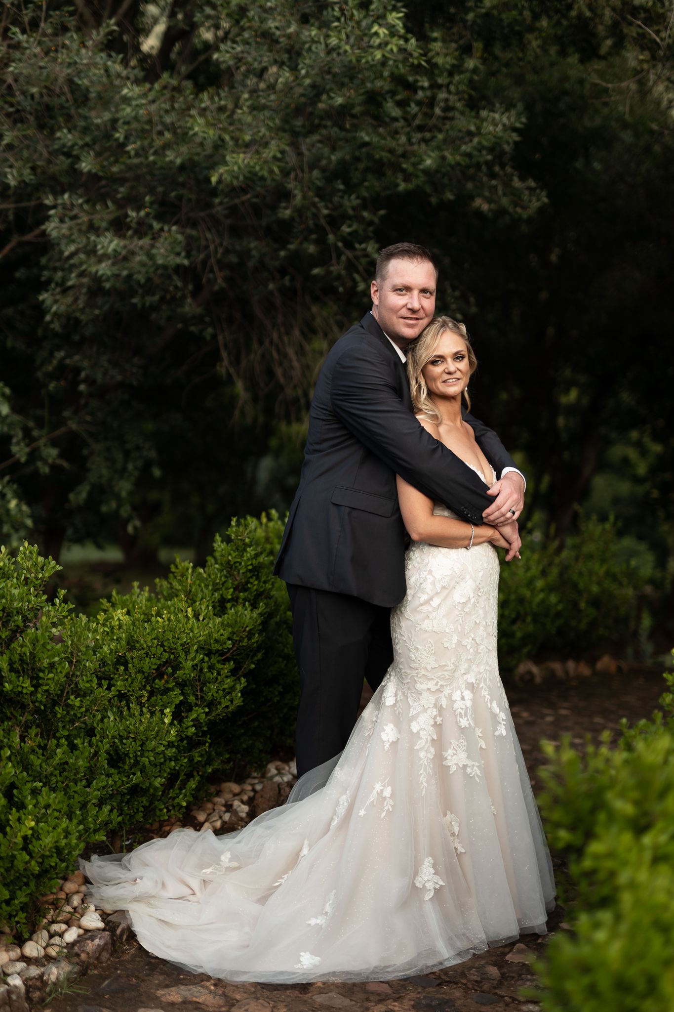A wedding couple poses on a stone path surrounded by manicured hedges and trees.