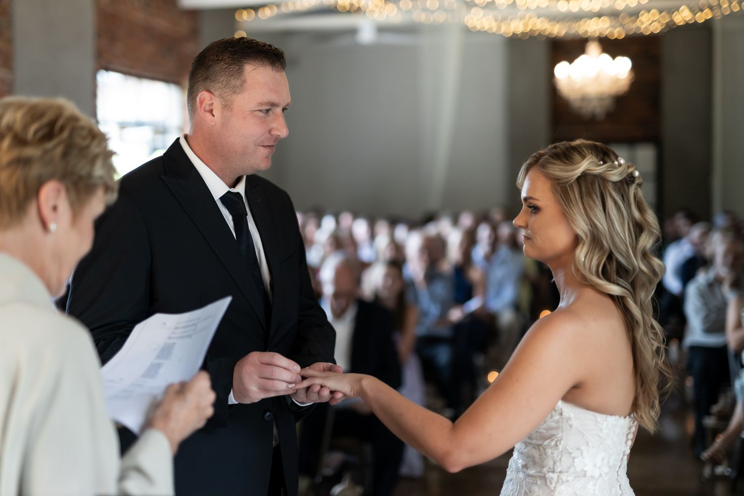 A couple exchanges rings during an indoor wedding ceremony with guests watching in the background.