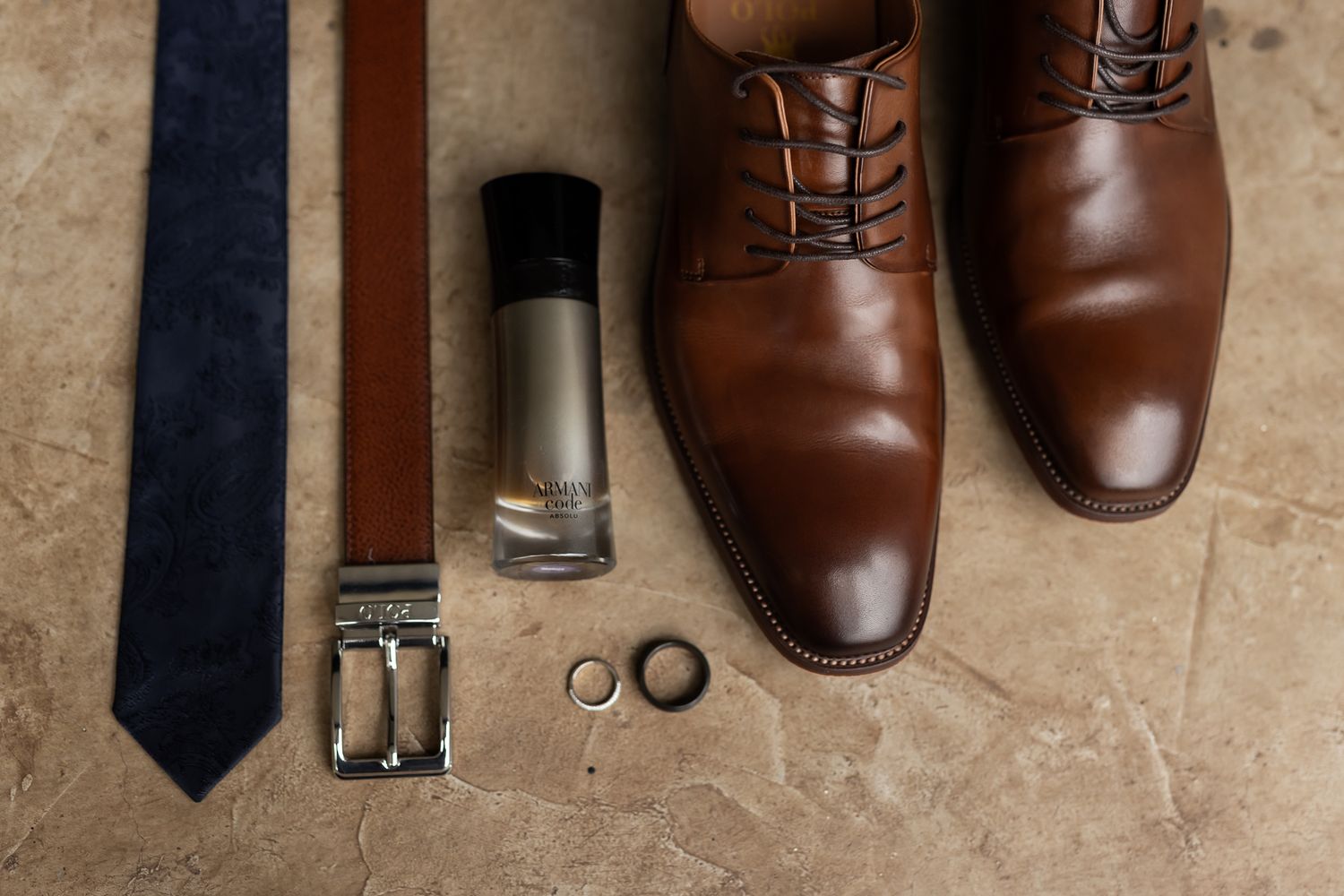 Brown leather dress shoes, navy tie and wedding rings arranged on beige floor.