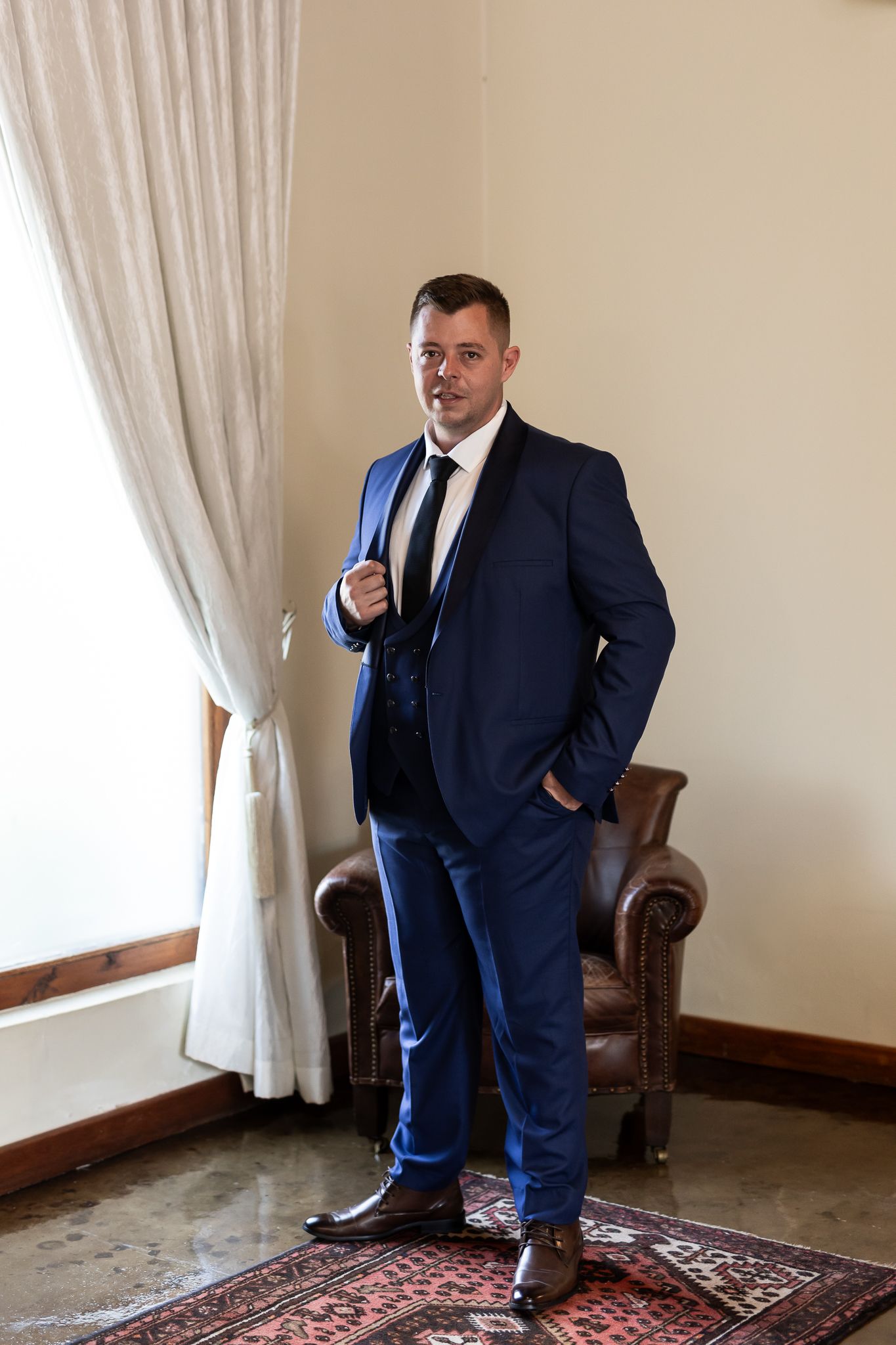 Full length portrait of person in navy suit standing by leather chair.
