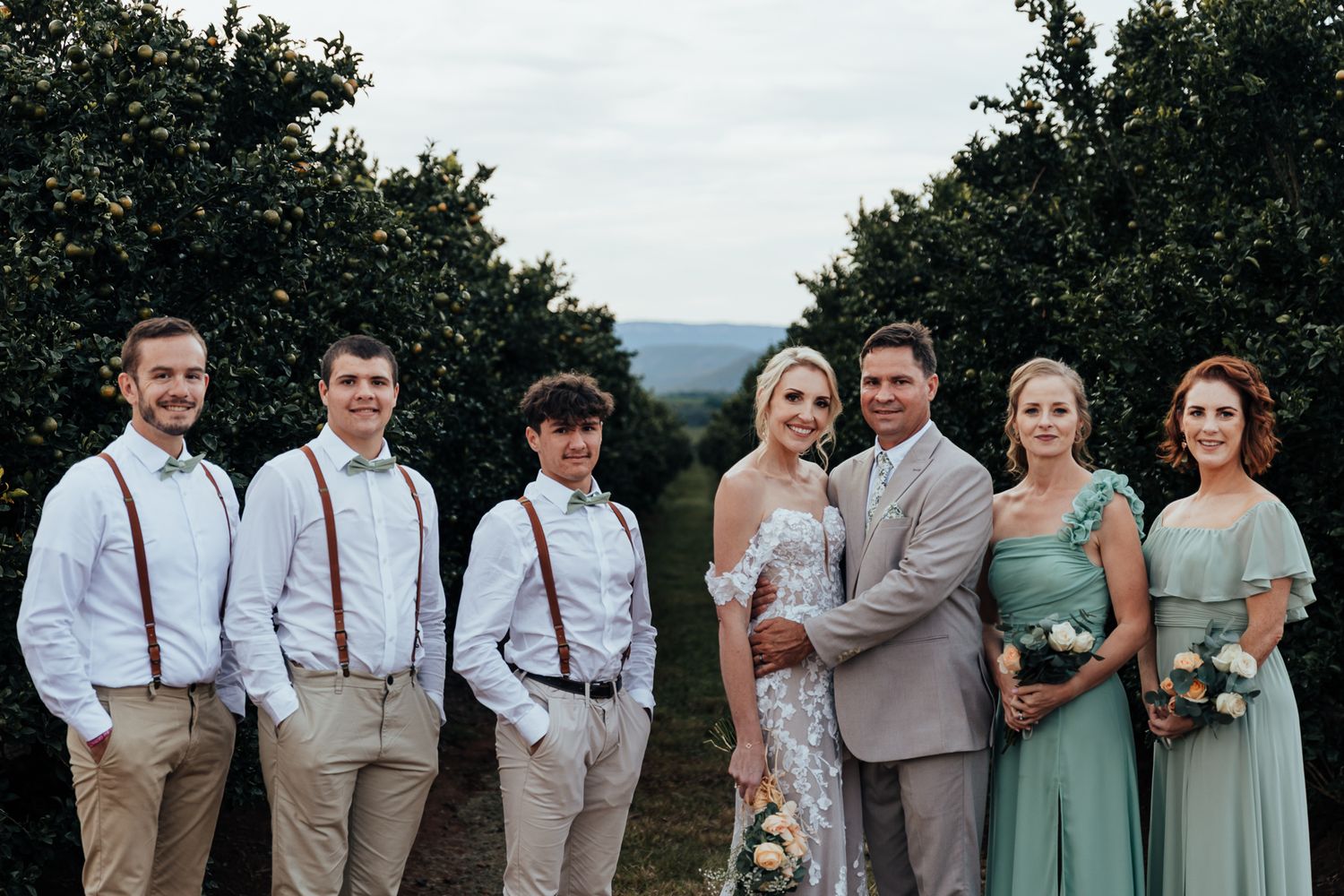 Wedding party in sage green dresses and suspenders poses together with mountain view backdrop.