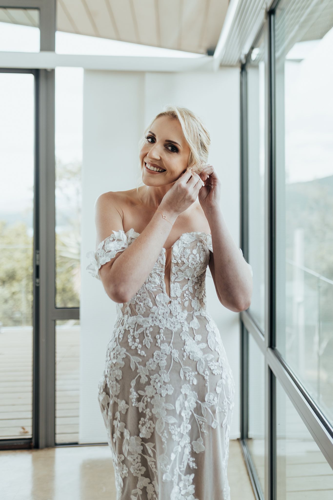 Joyful bridal portrait in detailed lace gown while putting on jewelry near bright windows.